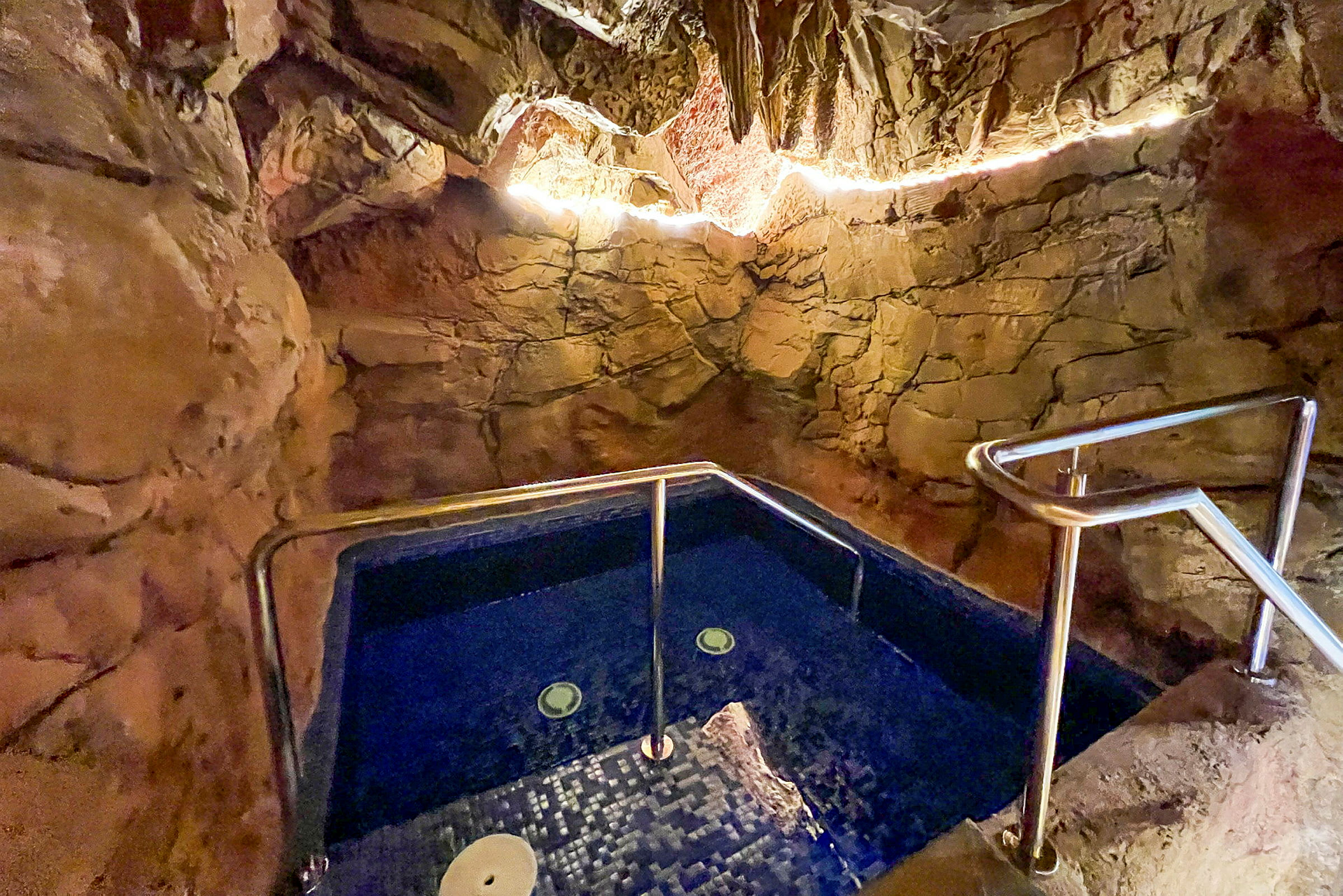 En jacuzzi omgiven av naturliknande stenväggar, med belysning längs taket. (AI-genererad text)