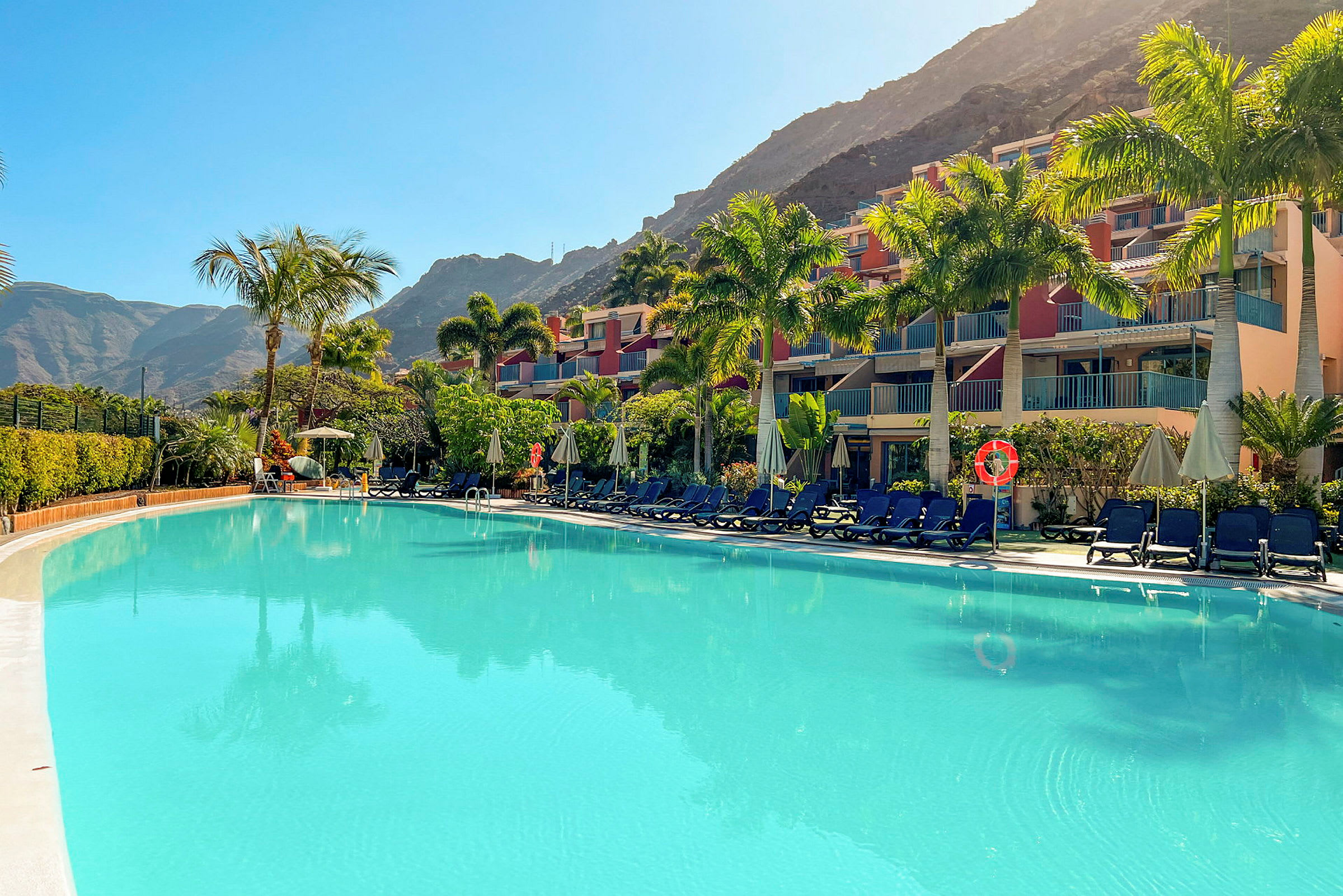 Bild av en swimmingpool, solstolar och hotellbyggnader omgivna av palmer vid foten av ett berg. (AI-genererad text)