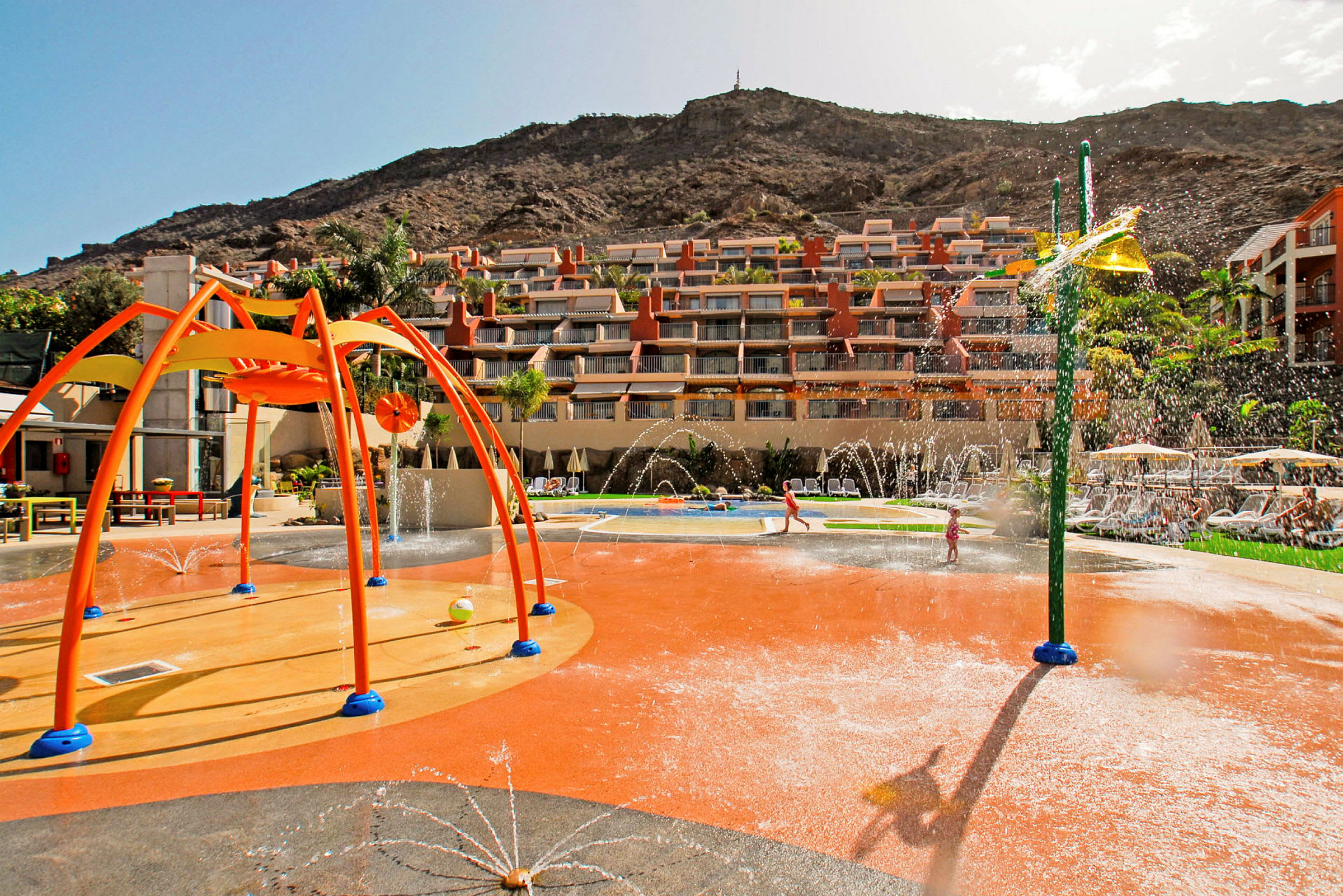 Lekplats med vattenfontäner vid hotellkomplex omgivet av berg. (AI-genererad text)