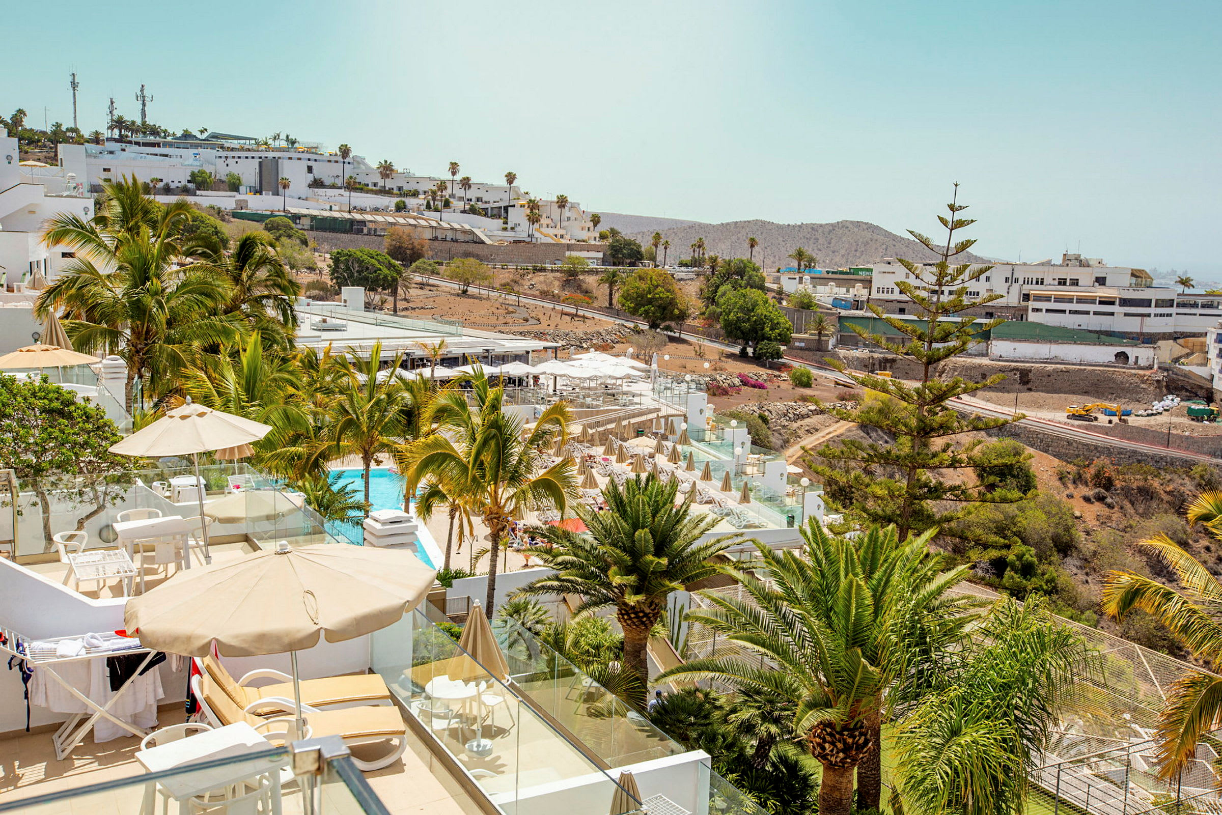 Vy över en hotellpool och terrass med solstolar och parasoller, omgivna av grönska och palmer. (AI-genererad text)