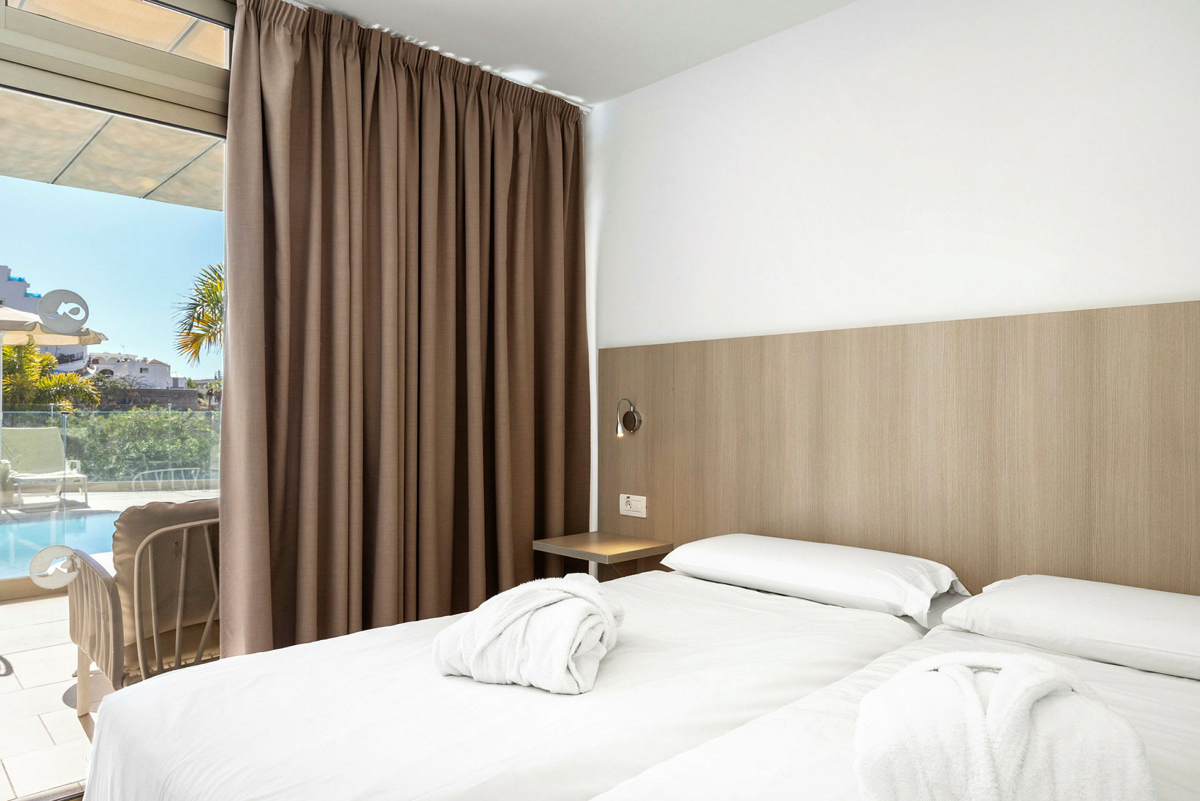 Ett hotellrum med två enkelsängar och vita lakan. Det finns bruna gardiner och en glasdörr som leder ut till en terrass med utsikt över en pool. (AI-genererad text)