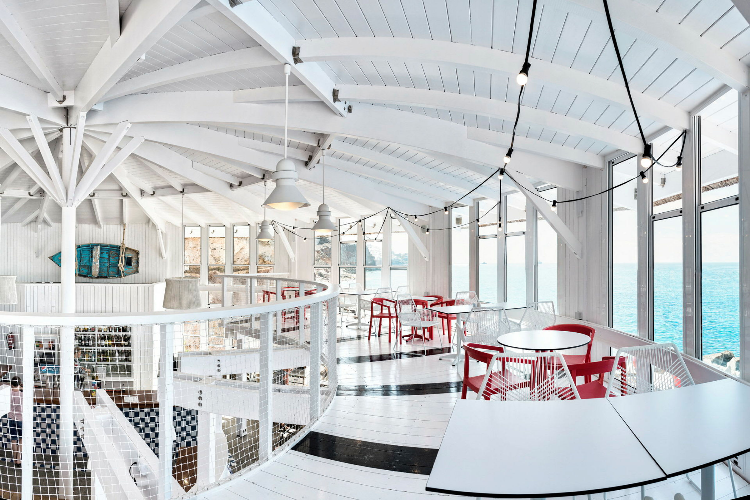 Ljust inredd restaurang med vita väggar, röda stolar och stora fönster med utsikt över havet. (AI-genererad text)