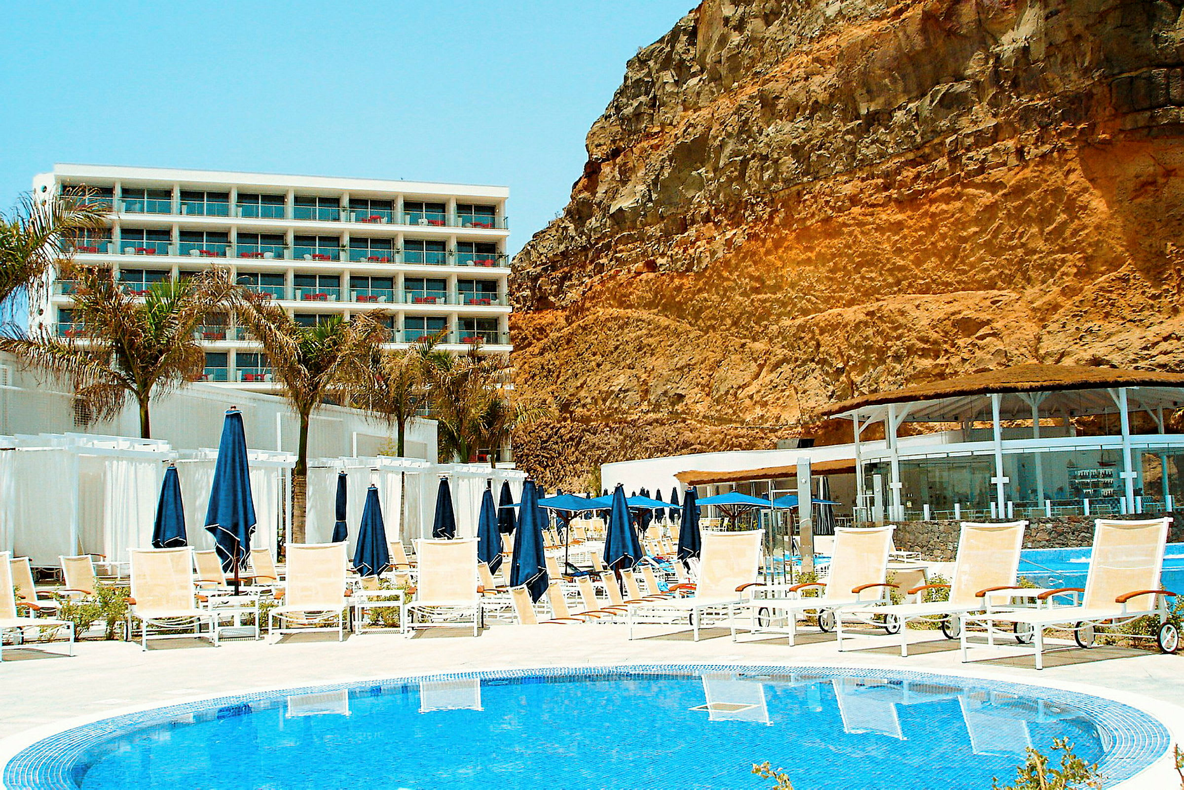 Bild av en hotellpool med solstolar och parasoller, med ett hotellkomplex i bakgrunden och en stor klippvägg vid sidan. (AI-genererad text)