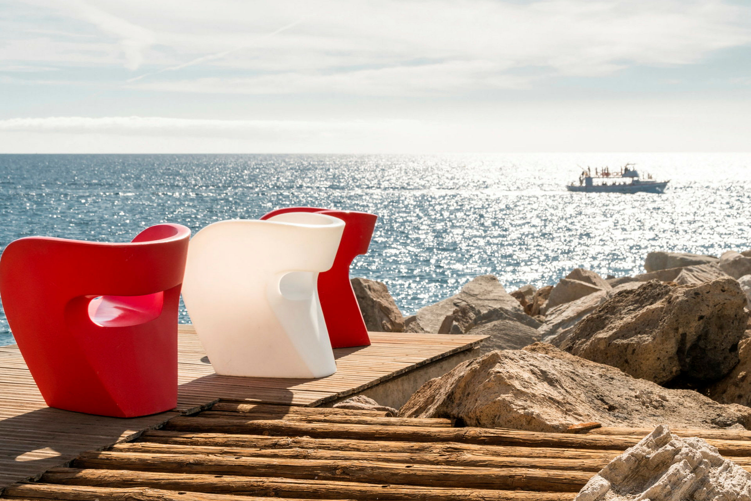Två moderna stolar, en röd och en vit, står på ett trädäck vid havet med en båt synlig i bakgrunden. (AI-genererad text)