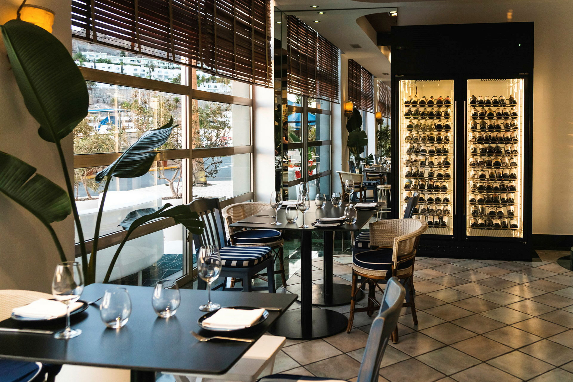 Interiör av restaurang med dukade bord, stora fönster mot utsikt och en upplyst vinförvaring. (AI-genererad text)