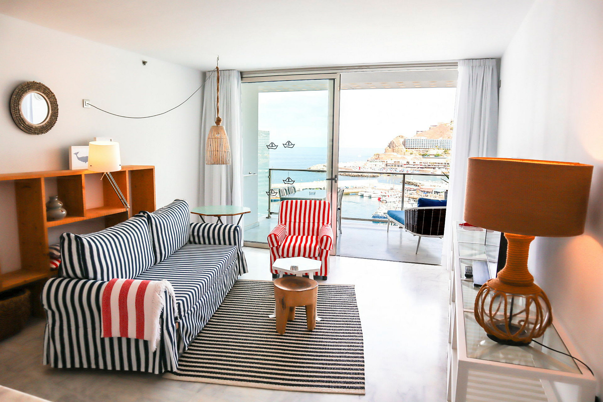 Ett hotellrum med maritim inredning, soffan har blå och vita ränder, fåtöljen har röda och vita ränder. Utsikt mot en hamn och hav från en balkong. (AI-genererad text)