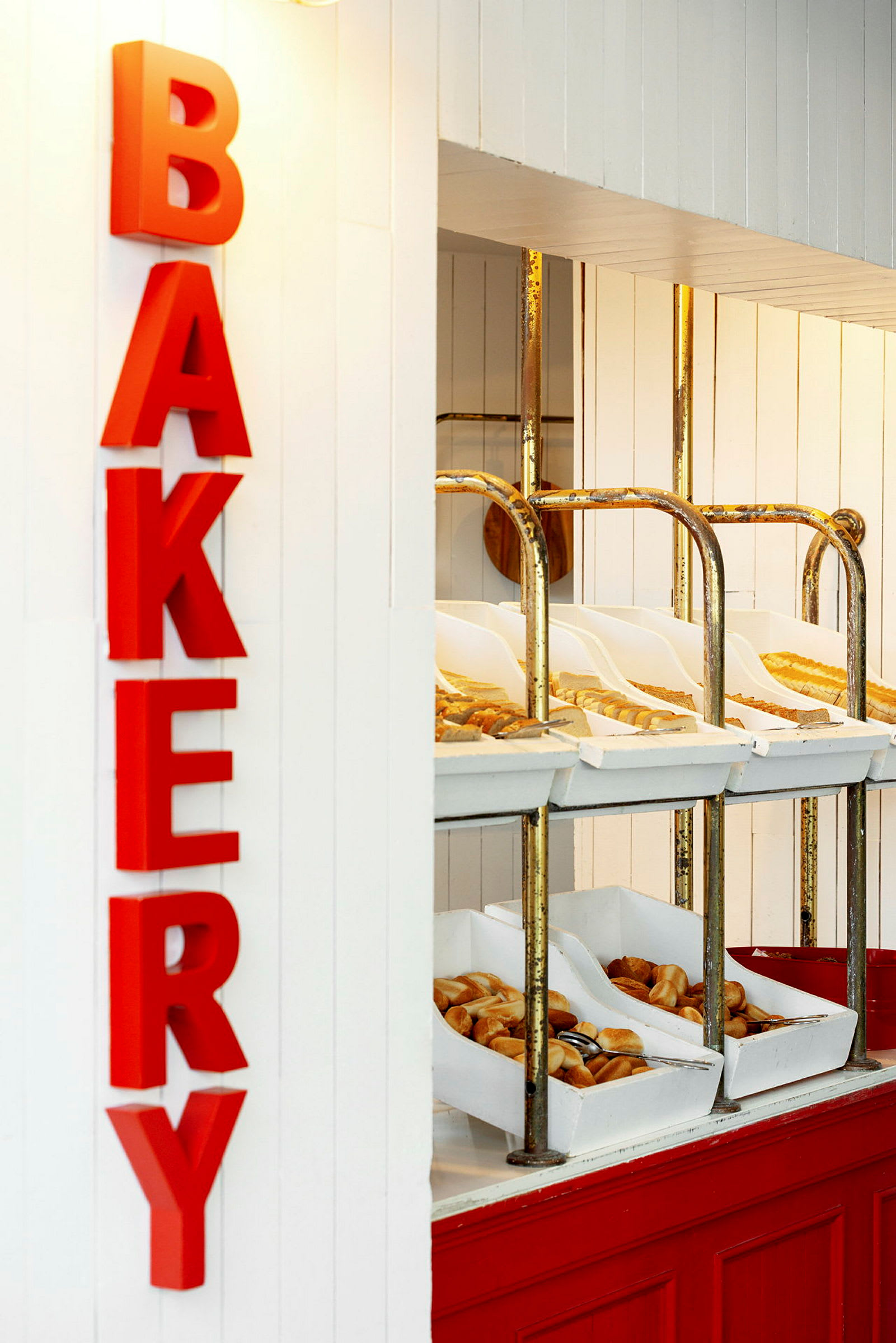 Ett bageriutrymme med röda bokstäver som stavar ordet 'BAKERY'. Hyllor innehåller olika typer av bröd och bakverk i vita lådor. (AI-genererad text)