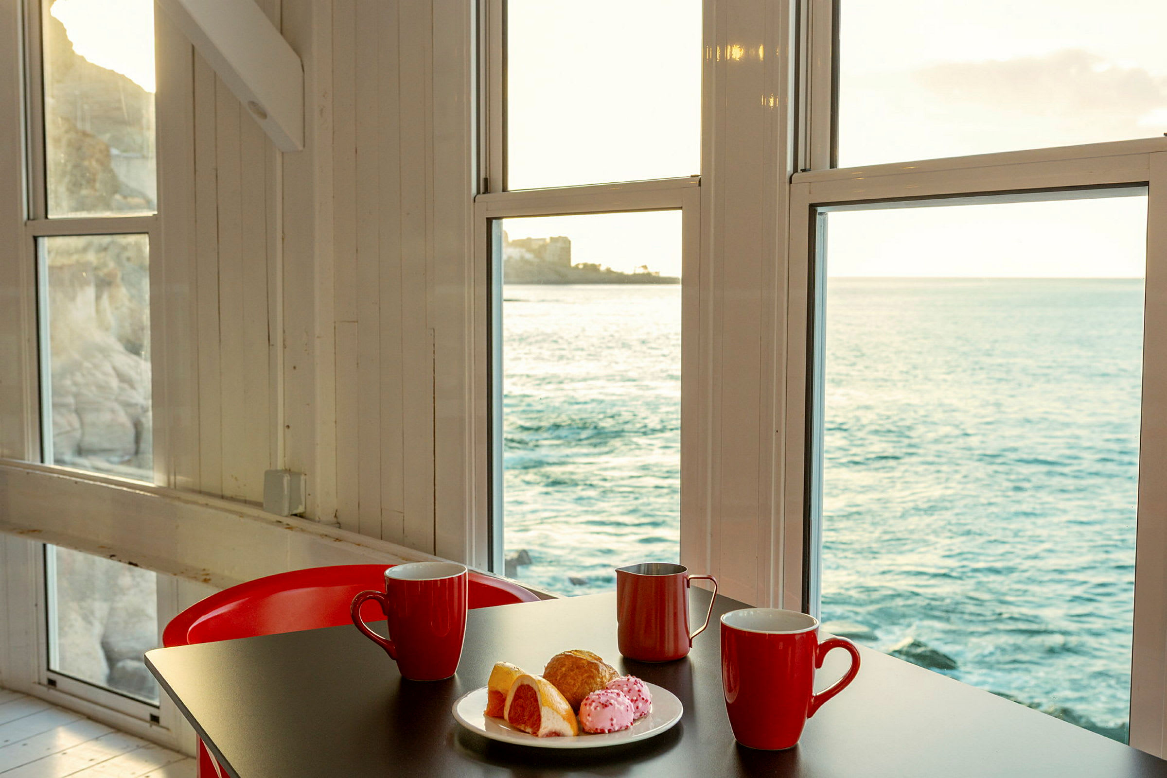 Ett bord med röda muggar och en tallrik med bakverk står framför stora fönster med utsikt över havet. (AI-genererad text)