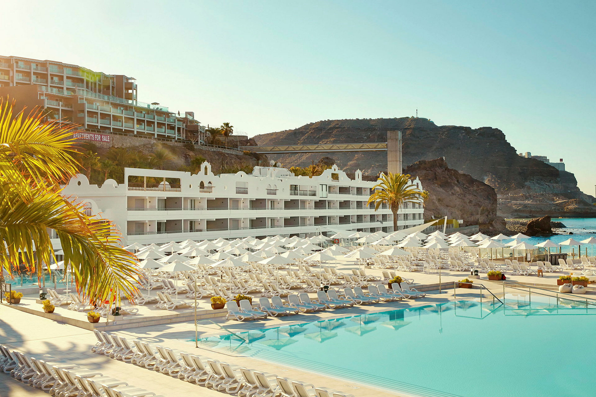 Hotellbyggnad med vita fasader, solstolar och parasoller vid en stor pool, samt klippformationer i bakgrunden. (AI-genererad text)