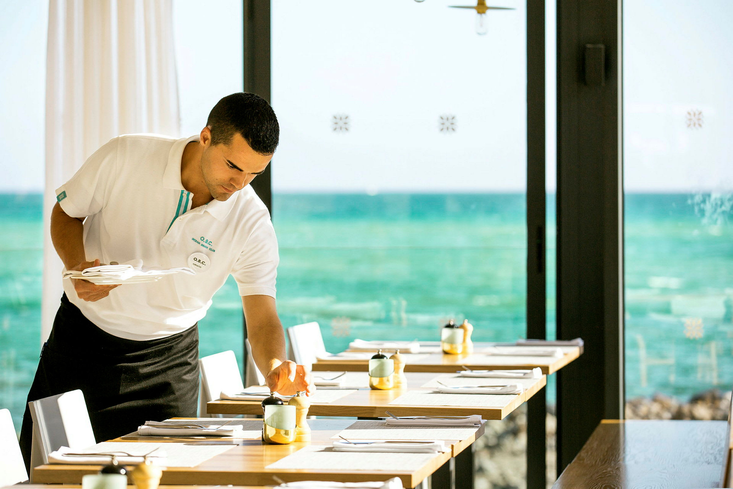 En servitör förbereder ett dukat bord med havet i bakgrunden. (AI-genererad text)