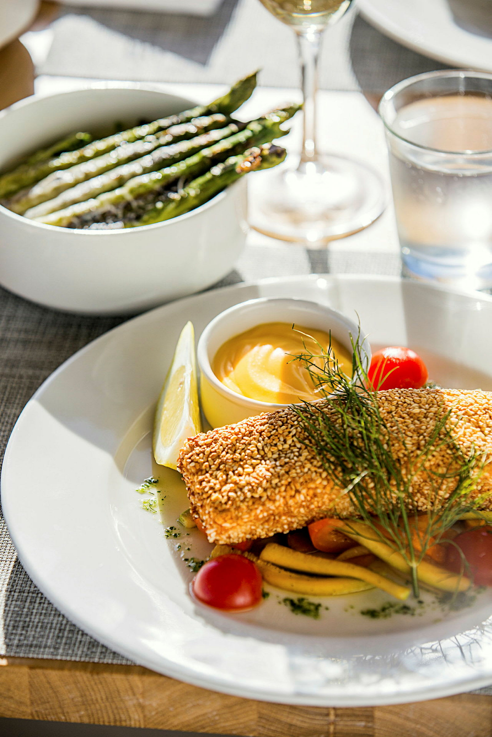 En tallrik med sesamöverbakad fisk, grönsaker och en skål med sås, samt en skål med grillad sparris bredvid. (AI-genererad text)