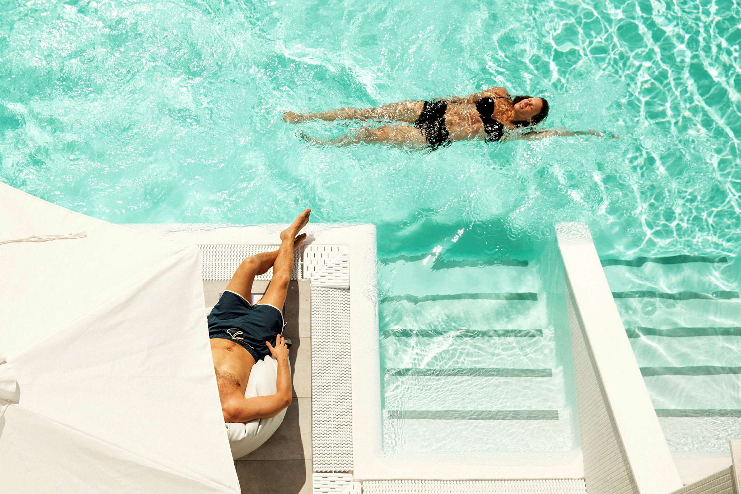 En kvinna simmar i en pool medan en man ligger och solar vid poolkanten. (AI-genererad text)