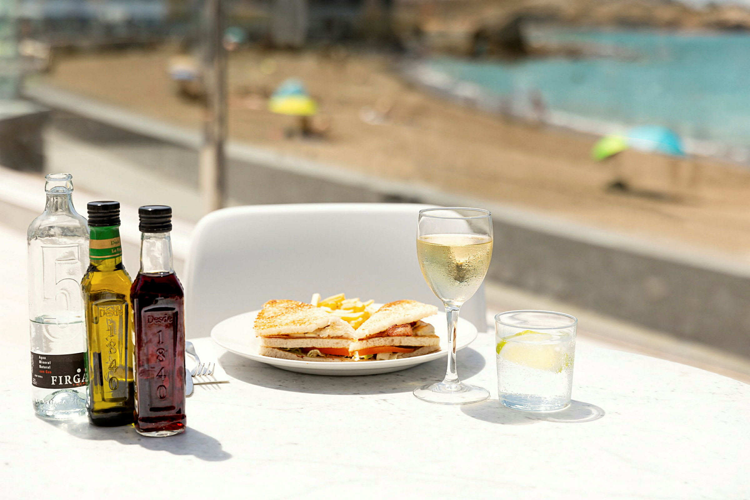 En tallrik med sandwich och pommes frites, omgiven av flaskor, ett glas vitt vin och ett glas vatten med lime, med en strand i bakgrunden. (AI-genererad text)