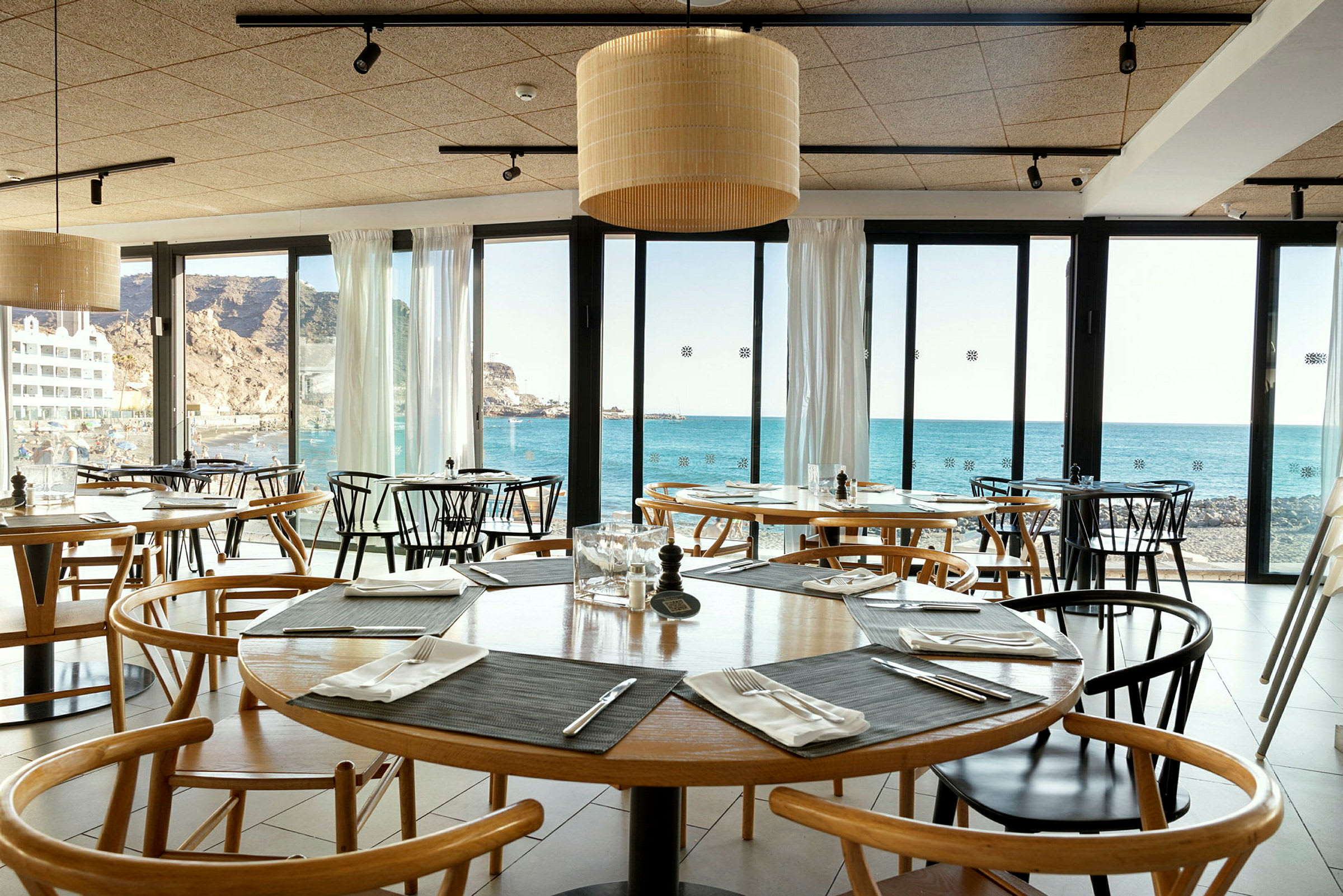 Insida av en restaurang med bord och stolar, utsikt mot havet genom glasfönster. (AI-genererad text)