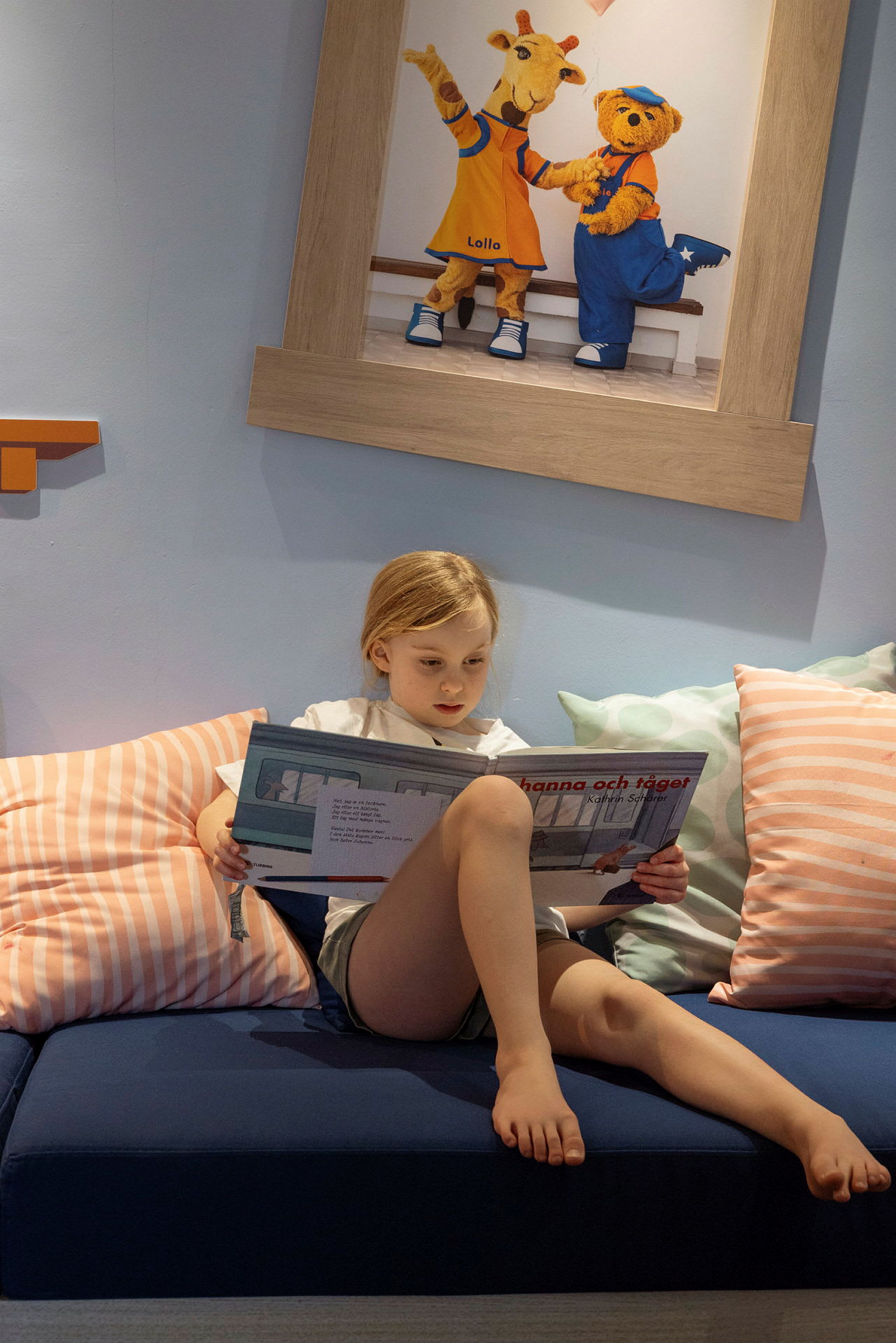 Ett barn sitter på en soffa och läser en bok, med en tavla föreställande djurkaraktärer i bakgrunden. (AI-genererad text)