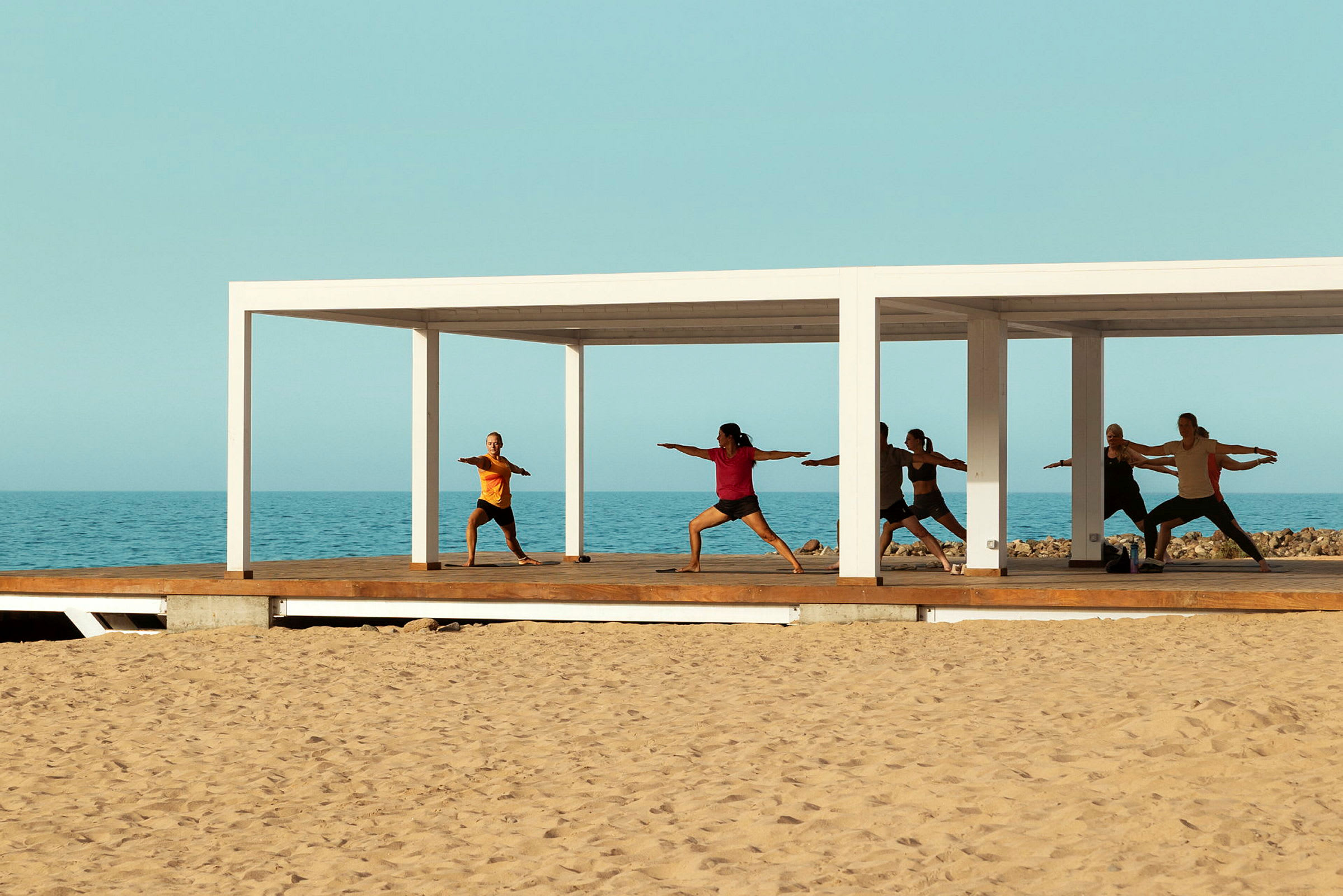 Personer som utför yoga på en träplattform vid havet under en vit pergola. (AI-genererad text)