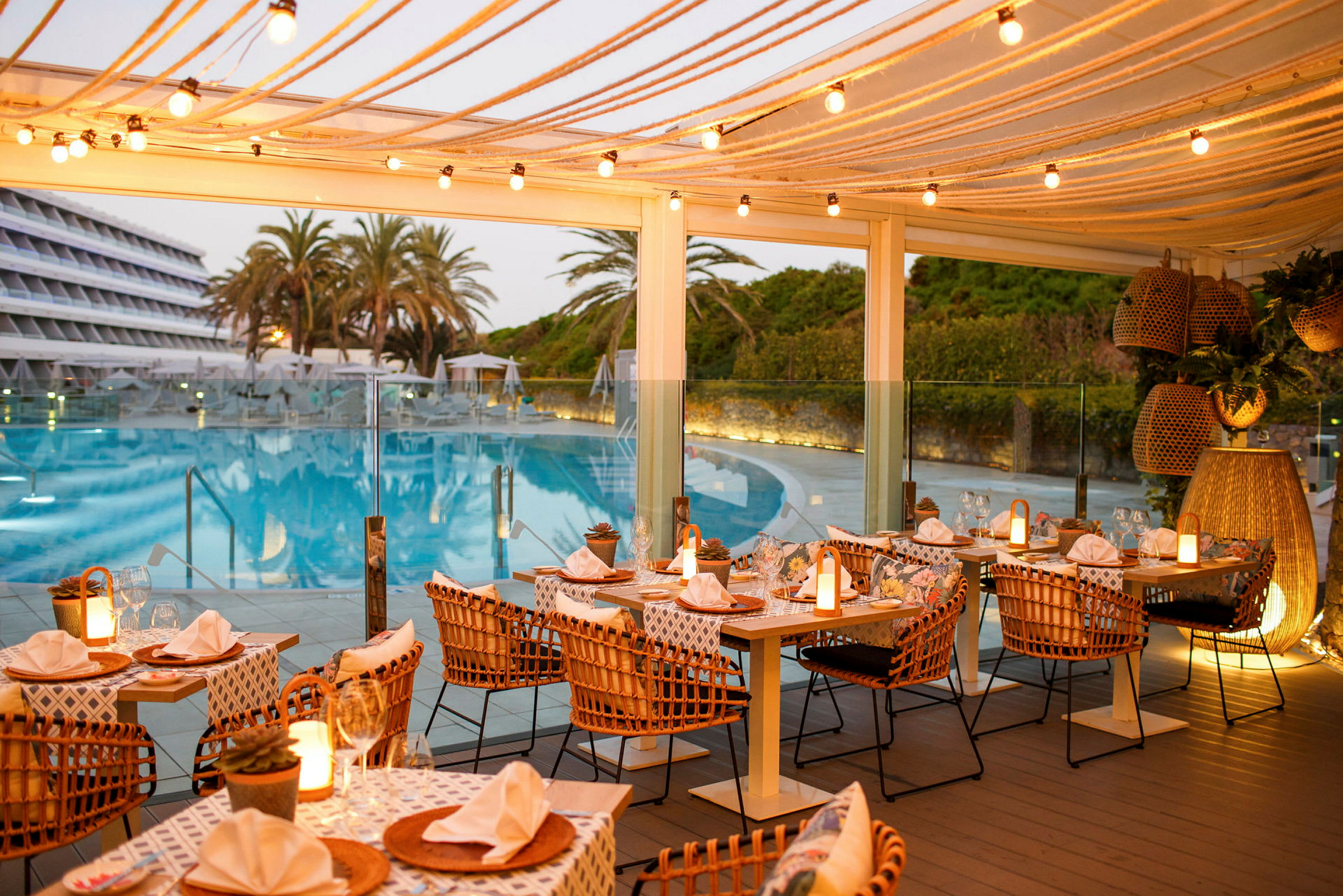 Utomhusrestaurang med dukade bord och ljusslingor, belägen nära en pool och omgiven av palmer och grönska. (AI-genererad text)