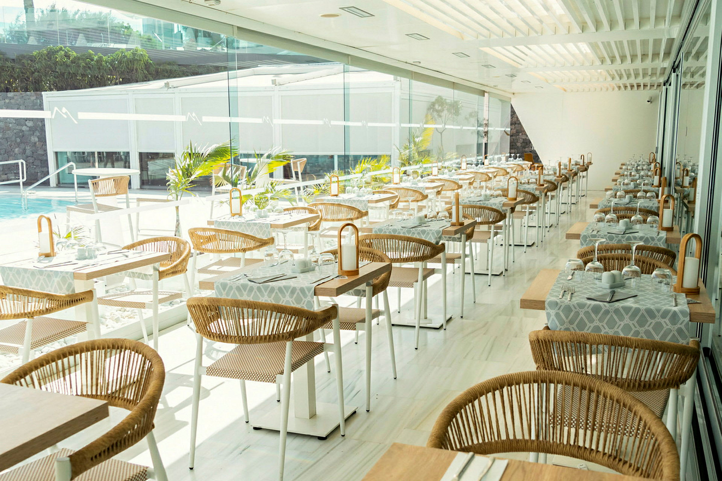 Modern restaurang med dukade bord och rottingstolar vid stora fönster. (AI-genererad text)