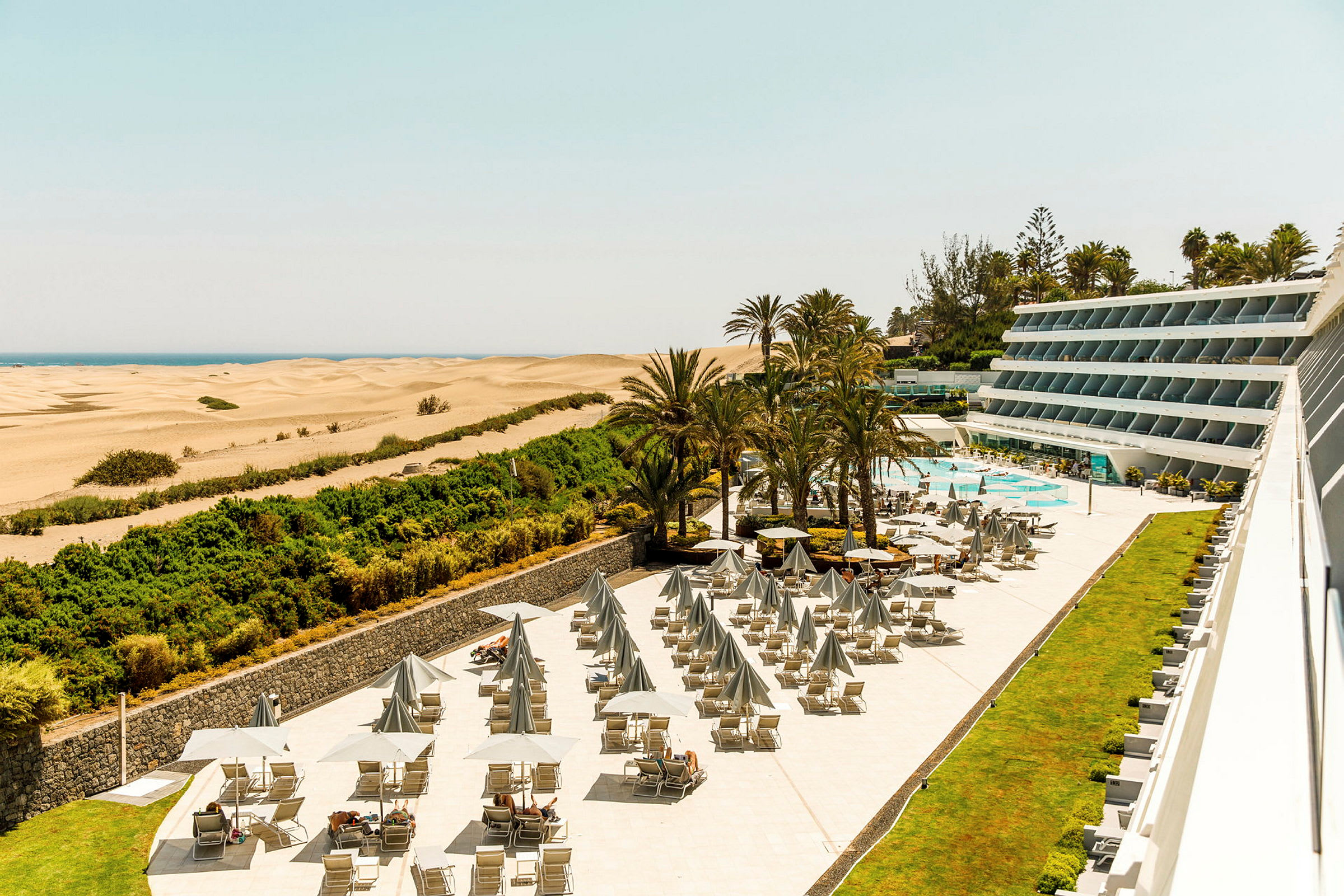 Utsikt över hotellområde med solstolar, parasoller och pool, intill sanddyner och hav. (AI-genererad text)