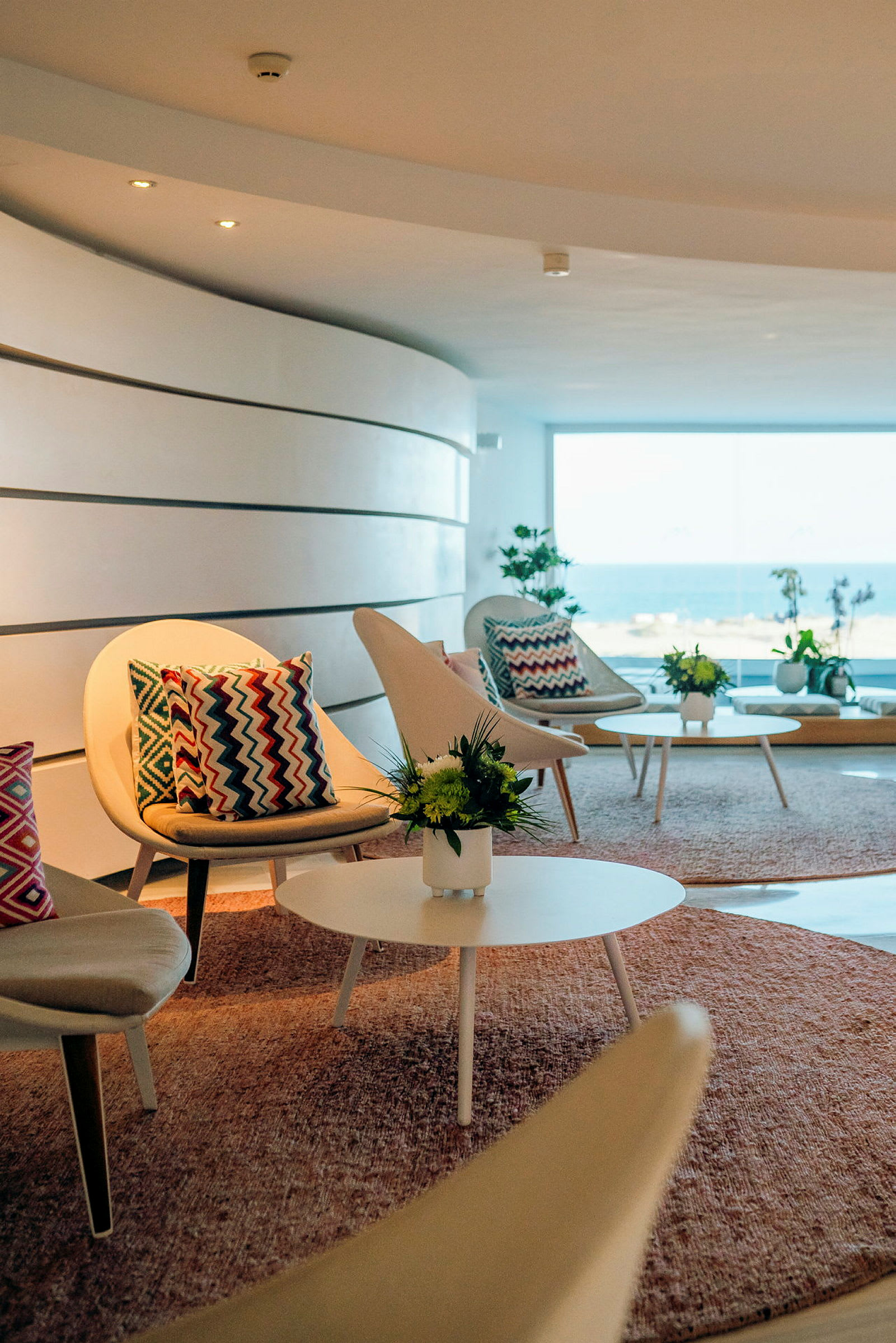 En lounge med moderna stolar och soffbord, dekorerade med färgglada kuddar och blommor, med havsutsikt genom ett stort fönster. (AI-genererad text)