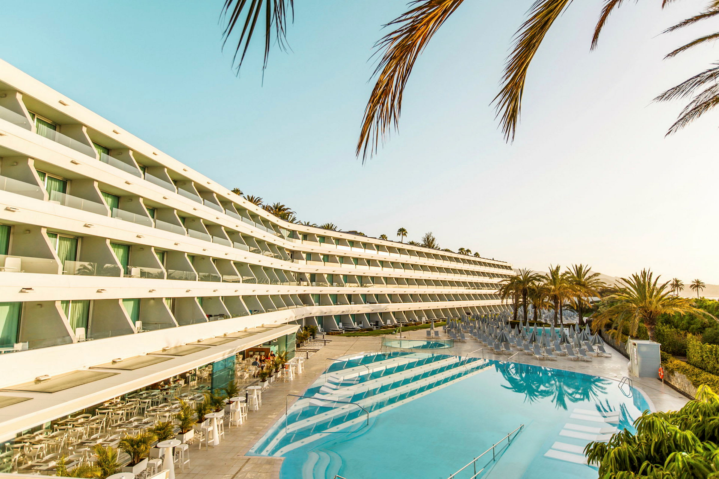 Modern hotellbyggnad med utsikt över en solig pool och omgivande palmer. (AI-genererad text)