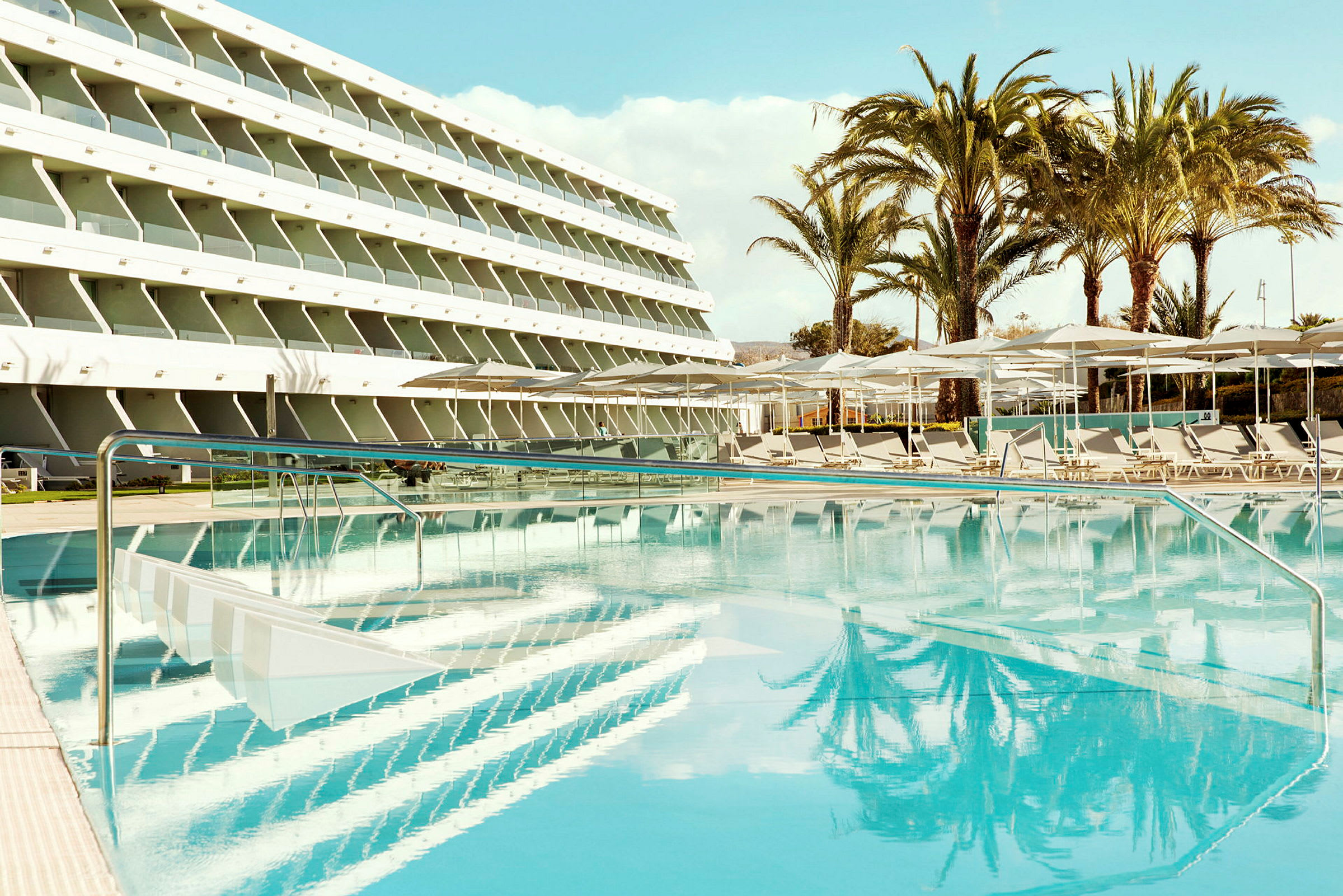 Poolområdet med solstolar, parasoller och ett fler våningshotell i bakgrunden. (AI-genererad text)