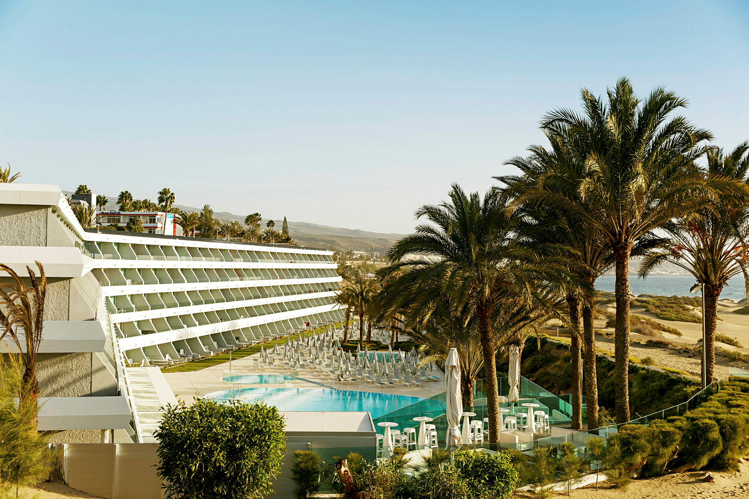 Hotellbyggnad med pool och solstolar omgiven av palmer vid stranden. (AI-genererad text)