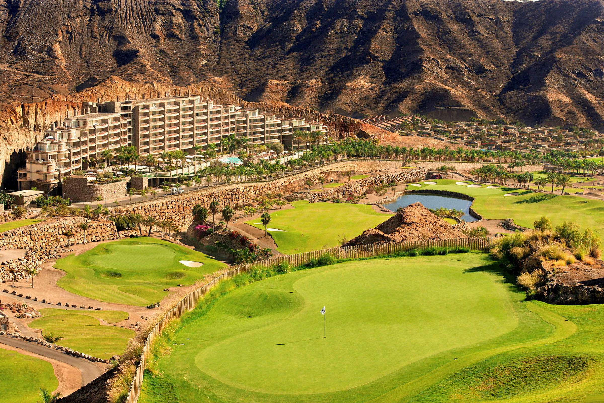 Hotellbyggnad omgiven av grönskande golfbanor och höga berg. (AI-genererad text)
