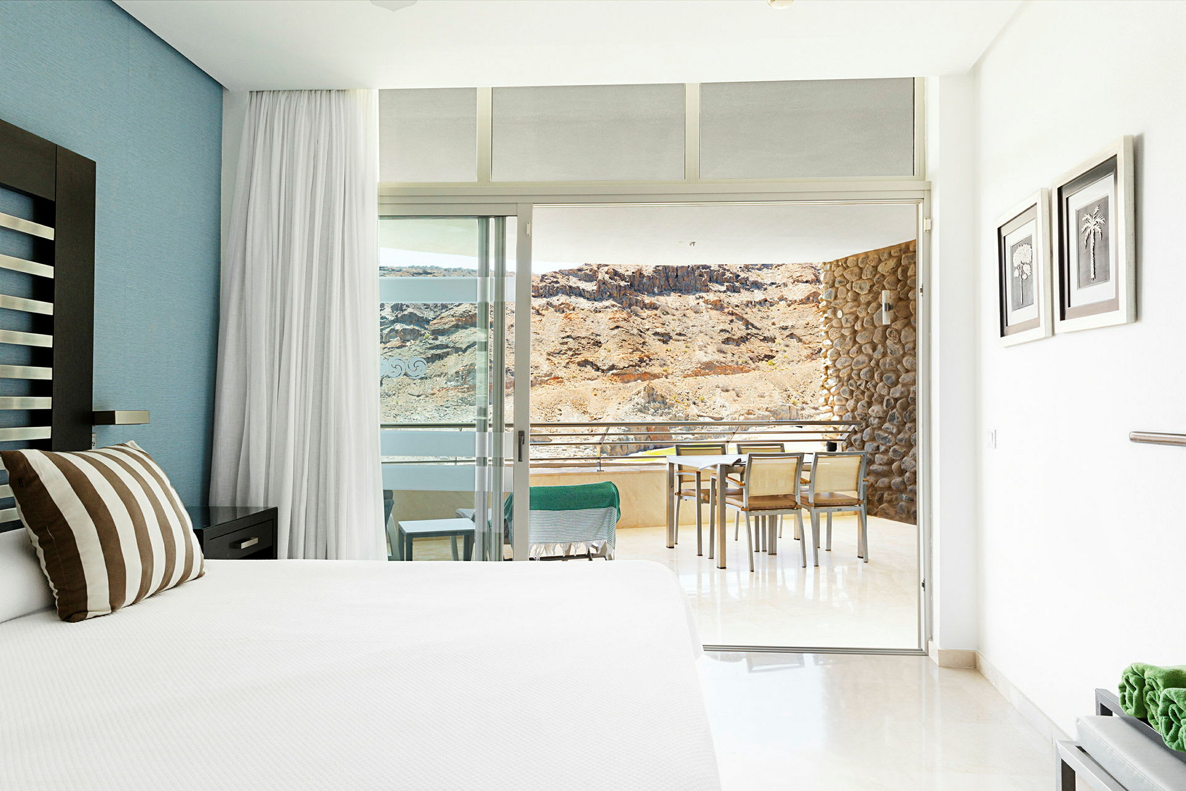 Hotellrum med en stor säng och utsikt mot en terrass med bord och stolar samt en bergig bakgrund. (AI-genererad text)