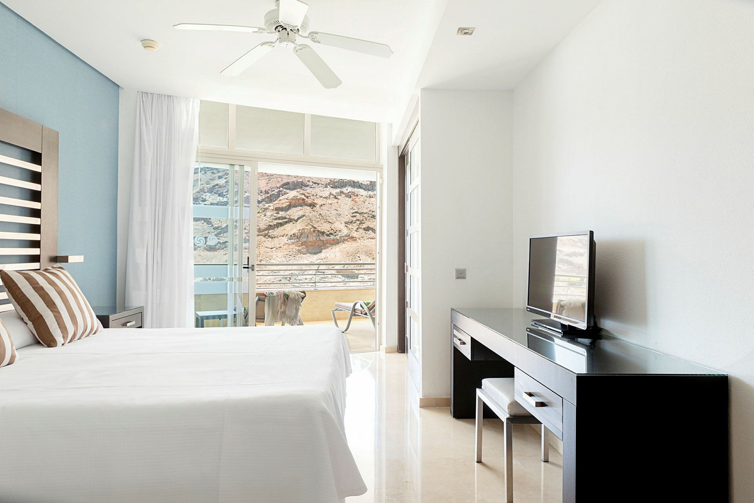 Ett hotellrum med en stor säng, TV och skrivbord samt en balkong med utsikt mot berg. (AI-genererad text)