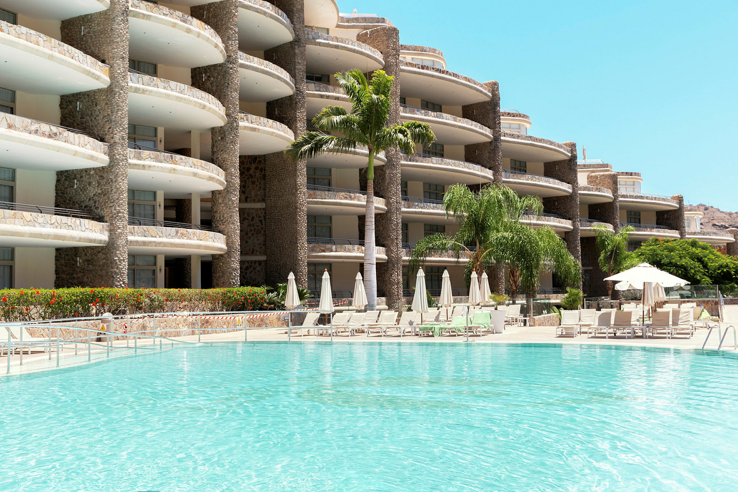 Bilden visar ett hotell med en pool framför och parasoller runtom. (AI-genererad text)