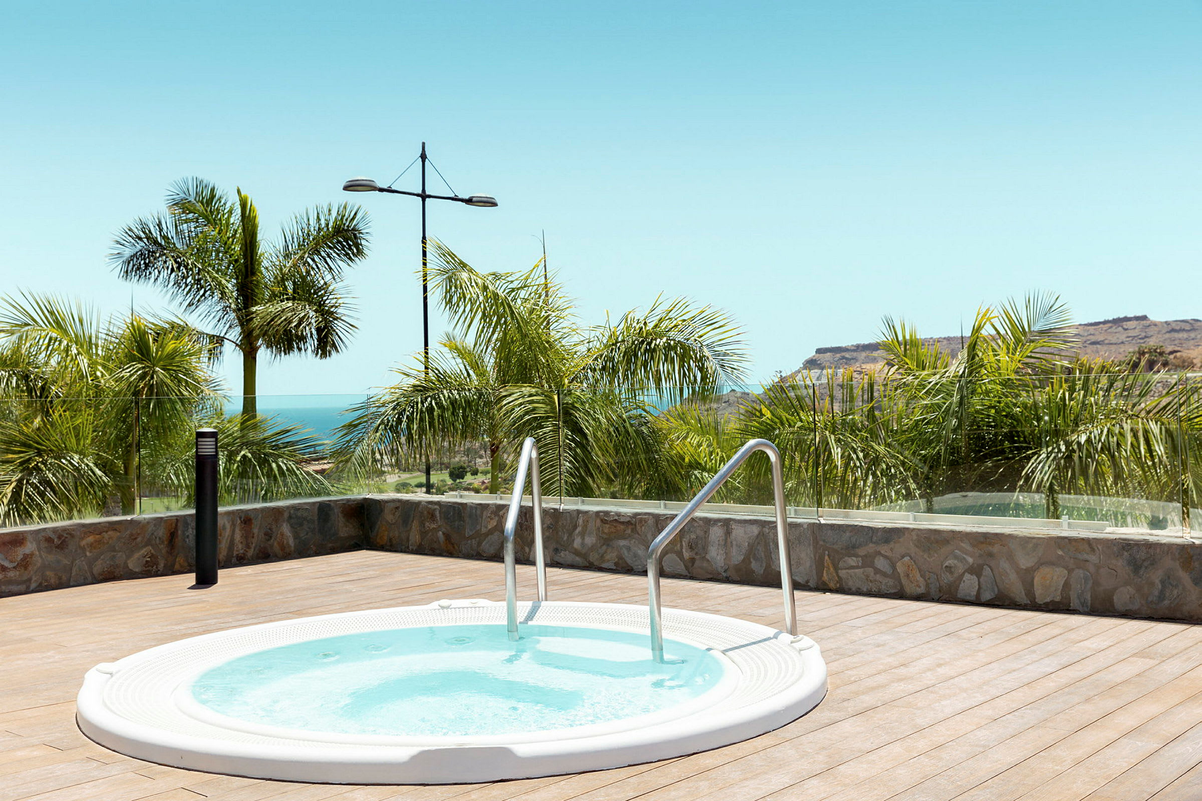 Rund jacuzzi på en terrass med havsutsikt och palmträd. (AI-genererad text)
