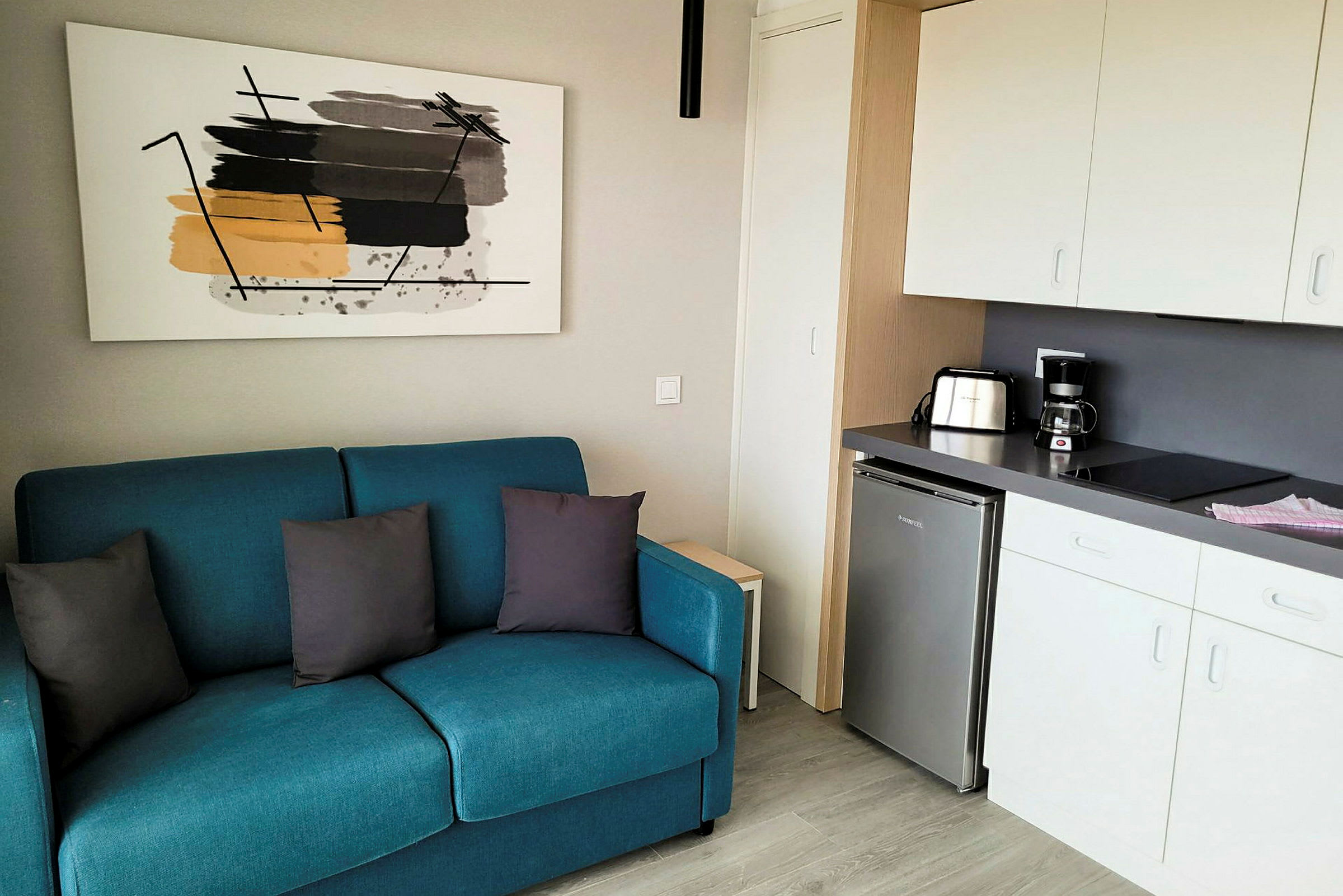 En liten lägenhet med en blå soffa, ett modernt kök och en abstrakt tavla på väggen. (AI-genererad text)