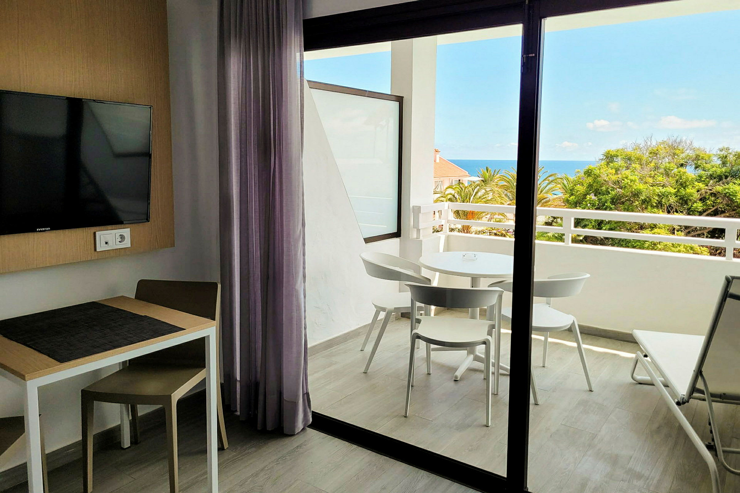 Ett hotellrum med bord, stol, en väggmonterad TV, och utsikt mot en balkong med möbler samt havet och grönska i bakgrunden. (AI-genererad text)