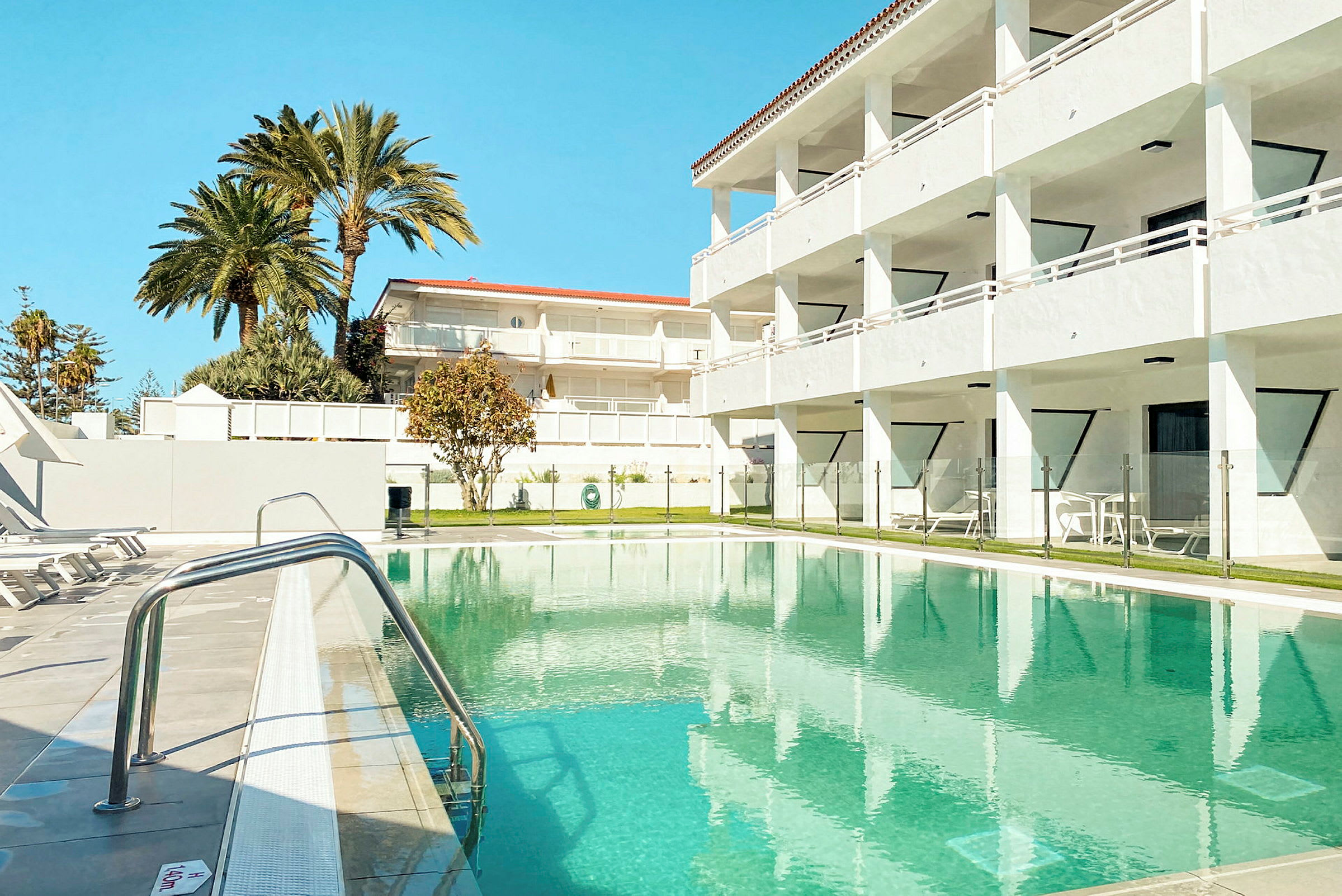 En hotellpool med solstolar vid sidan, balkonhus och palmer i bakgrunden. (AI-genererad text)
