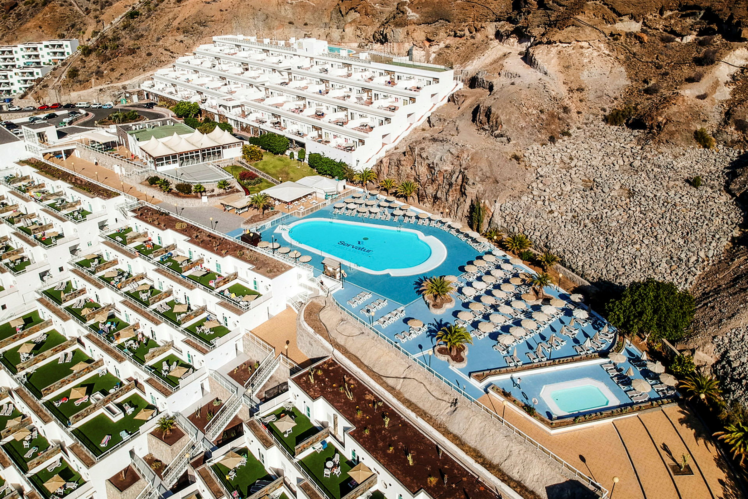 Flygbild av ett hotellområde med vita byggnader, gröna uteplatser, en stor pool och omgivande berg. (AI-genererad text)
