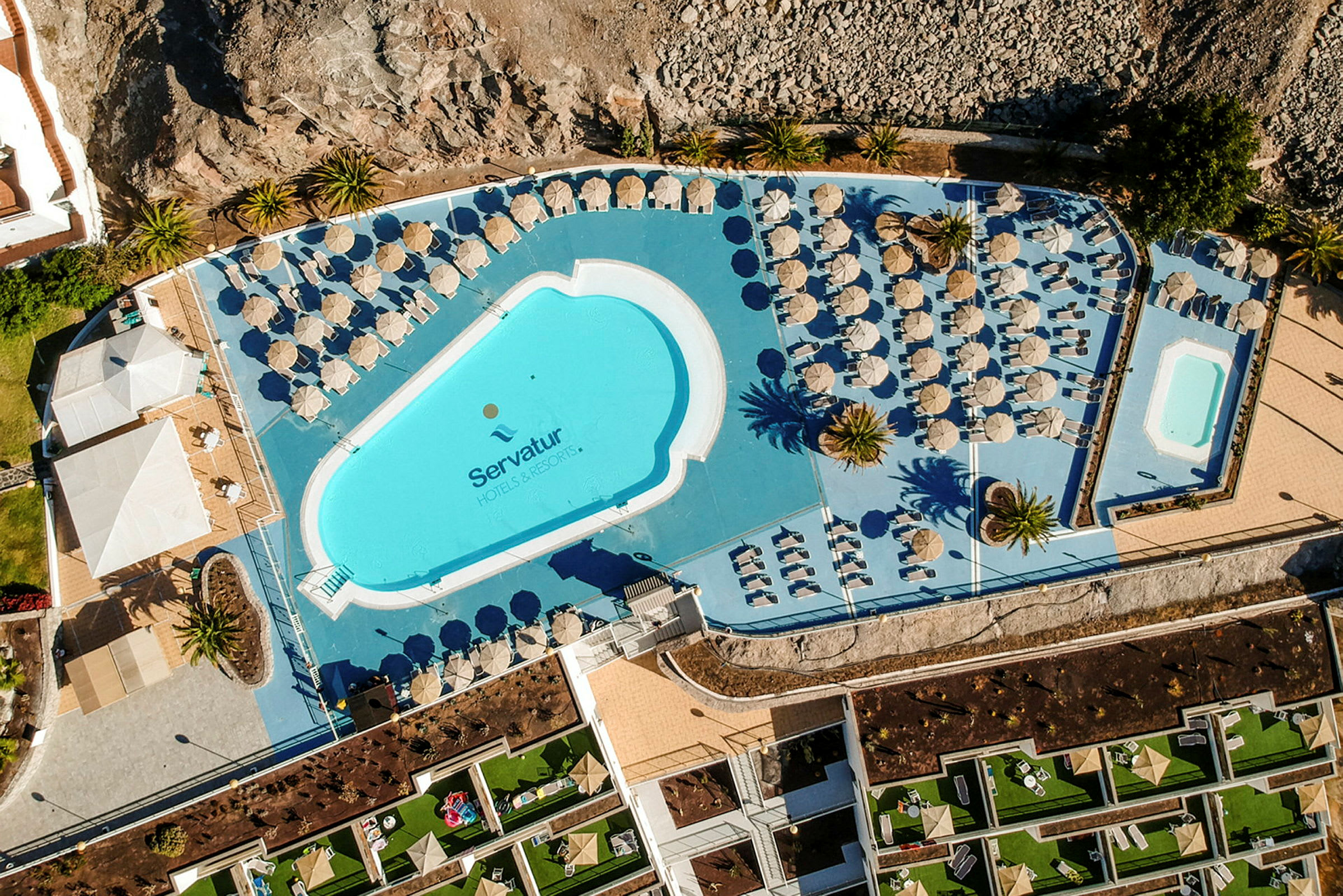 Luftbild av poolområdet på ett hotell med solstolar och parasoller. (AI-genererad text)