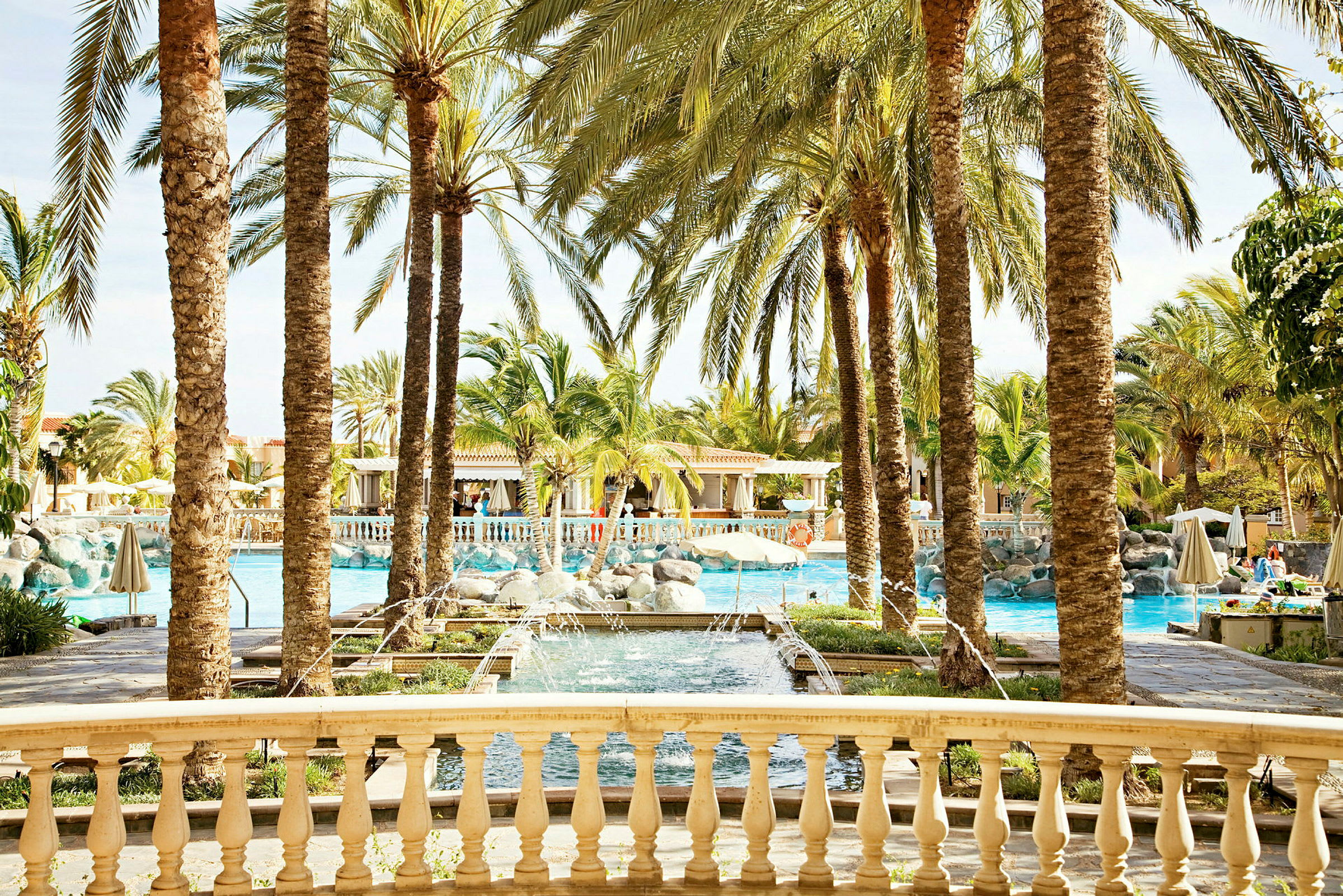 Utsikt över en pool omgiven av palmer och en stenbro vid ett semesterresort. (AI-genererad text)