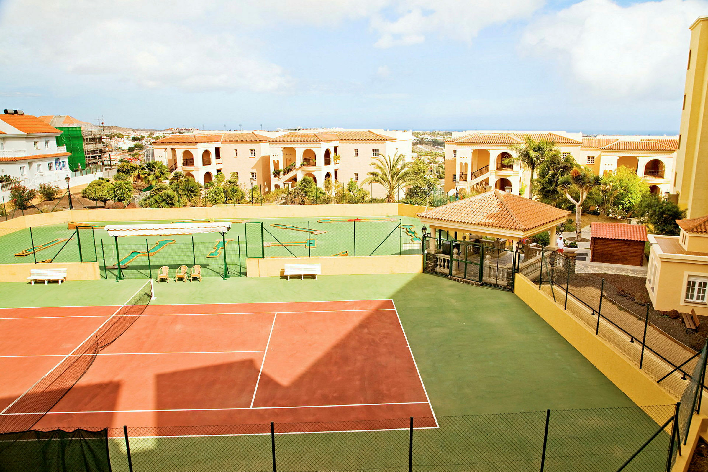 En tennisbana med en minigolfanläggning i bakgrunden omgiven av byggnader. (AI-genererad text)