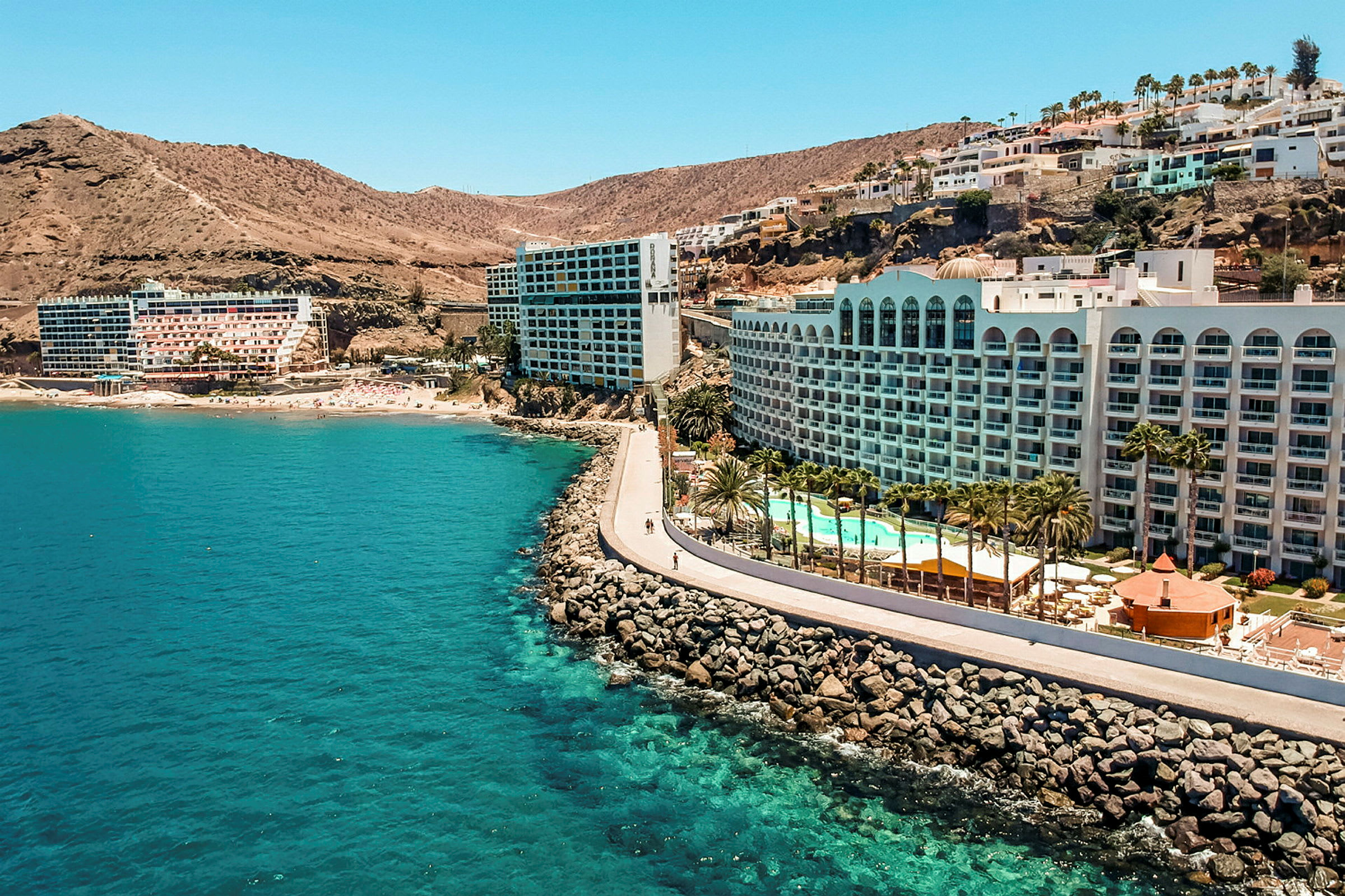 Hotell vid en kust med klippor, pool och palmer. (AI-genererad text)