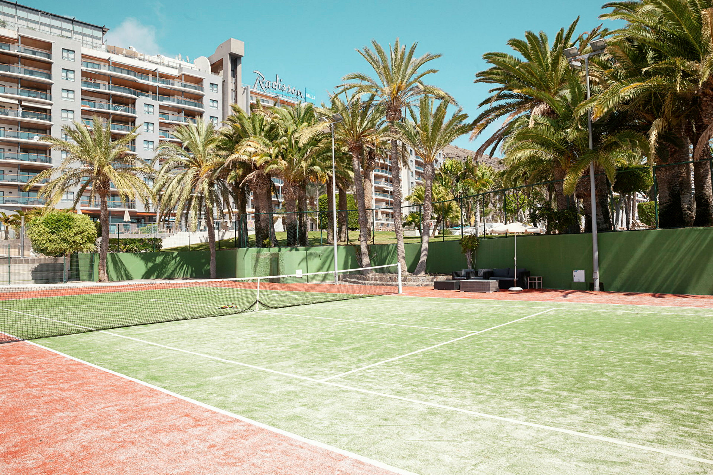 Tennisplan med grusunderlag omgiven av palmer, med ett hotell i bakgrunden. (AI-genererad text)
