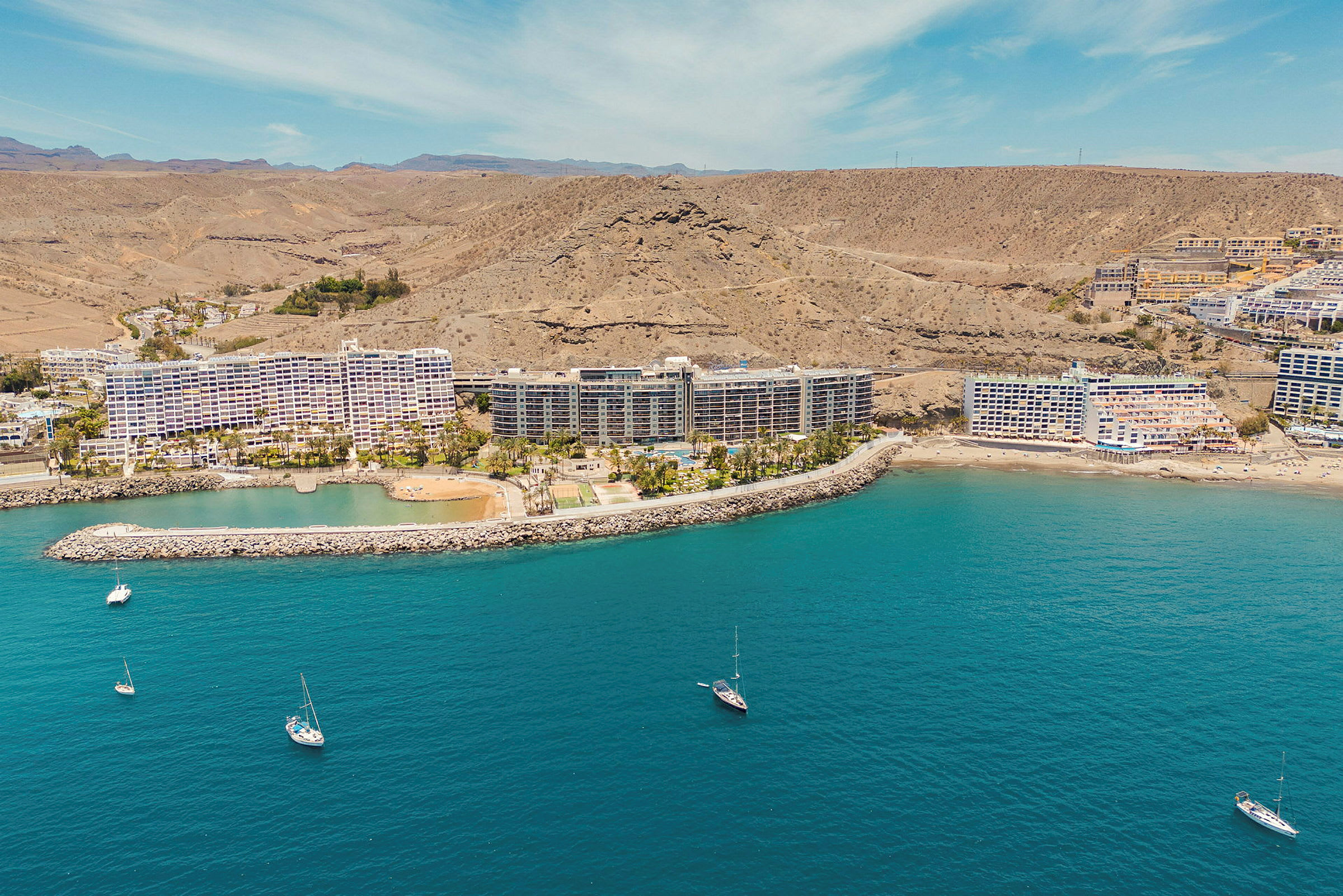 Flygfoto av kusten med hotellbyggnader, småbåtar och berg i bakgrunden. (AI-genererad text)