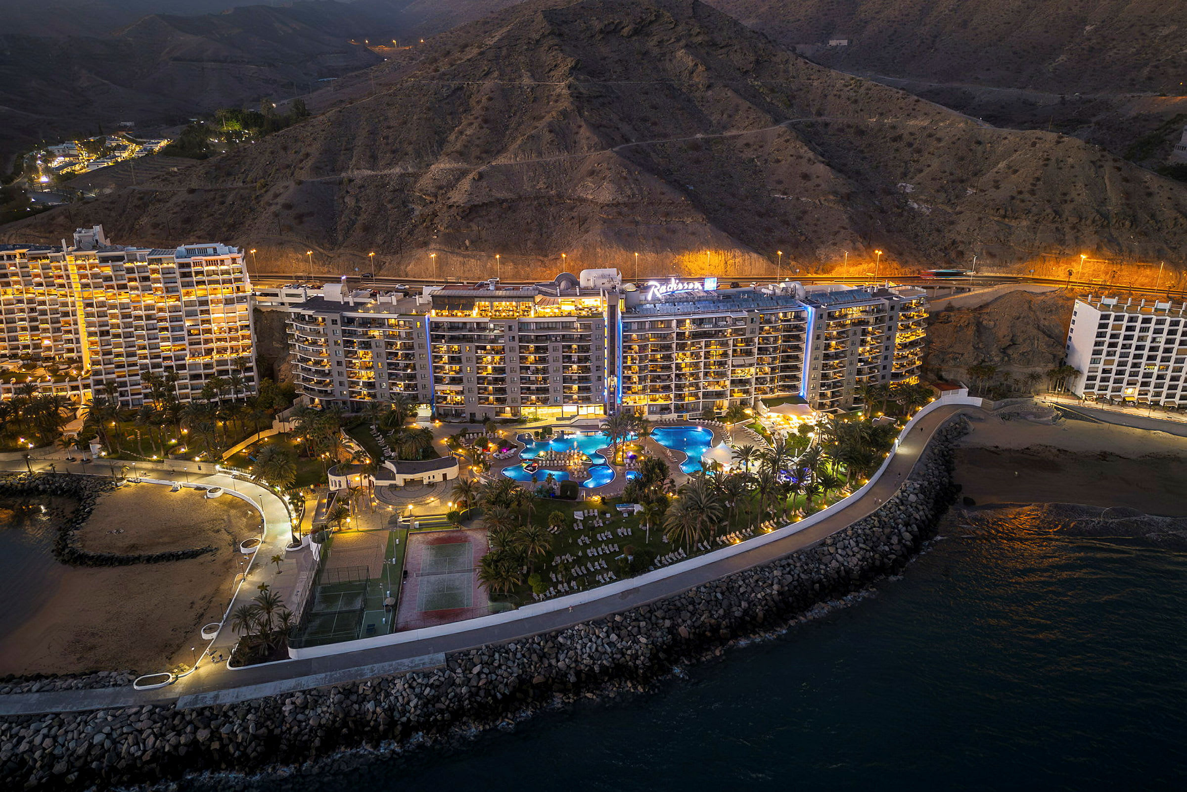 Flygbild av ett hotellkomplex med poolområde nära kusten, berg i bakgrunden. (AI-genererad text)