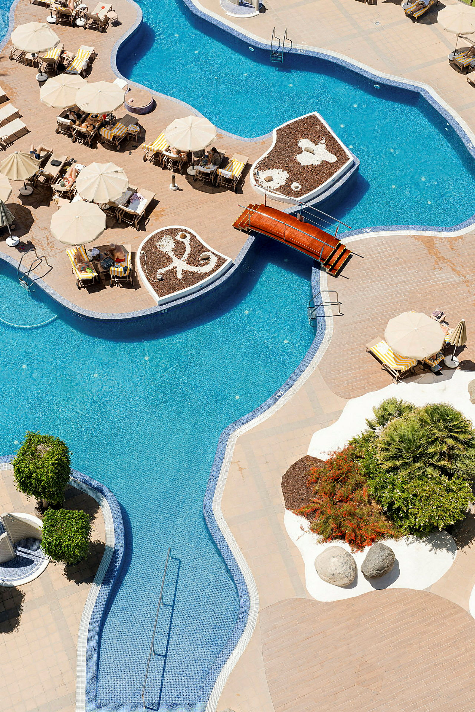 Bild av en slingrande swimmingpool med solstolar och parasoller intill, samt dekorativa planteringar och gångbro. (AI-genererad text)