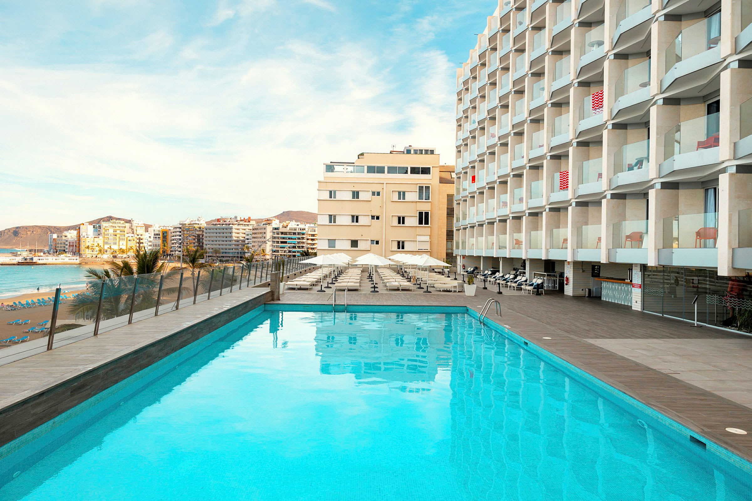 Utomhuspool framför hotellbyggnad, med strand och stad i bakgrunden. (AI-genererad text)