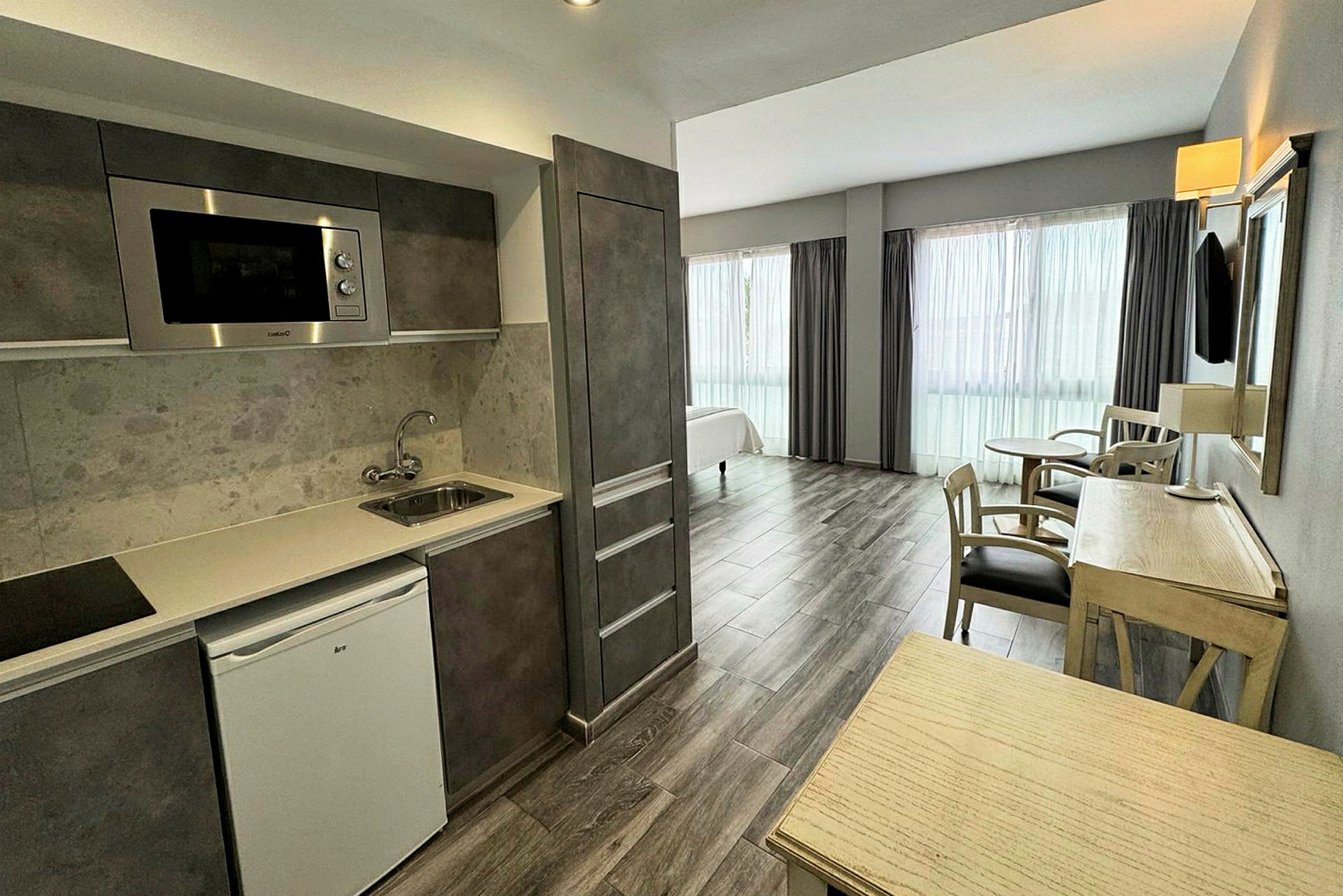 Ett hotellrum med integrerat kök och vardagsområde, möblerat med träbord och stolar. (AI-genererad text)