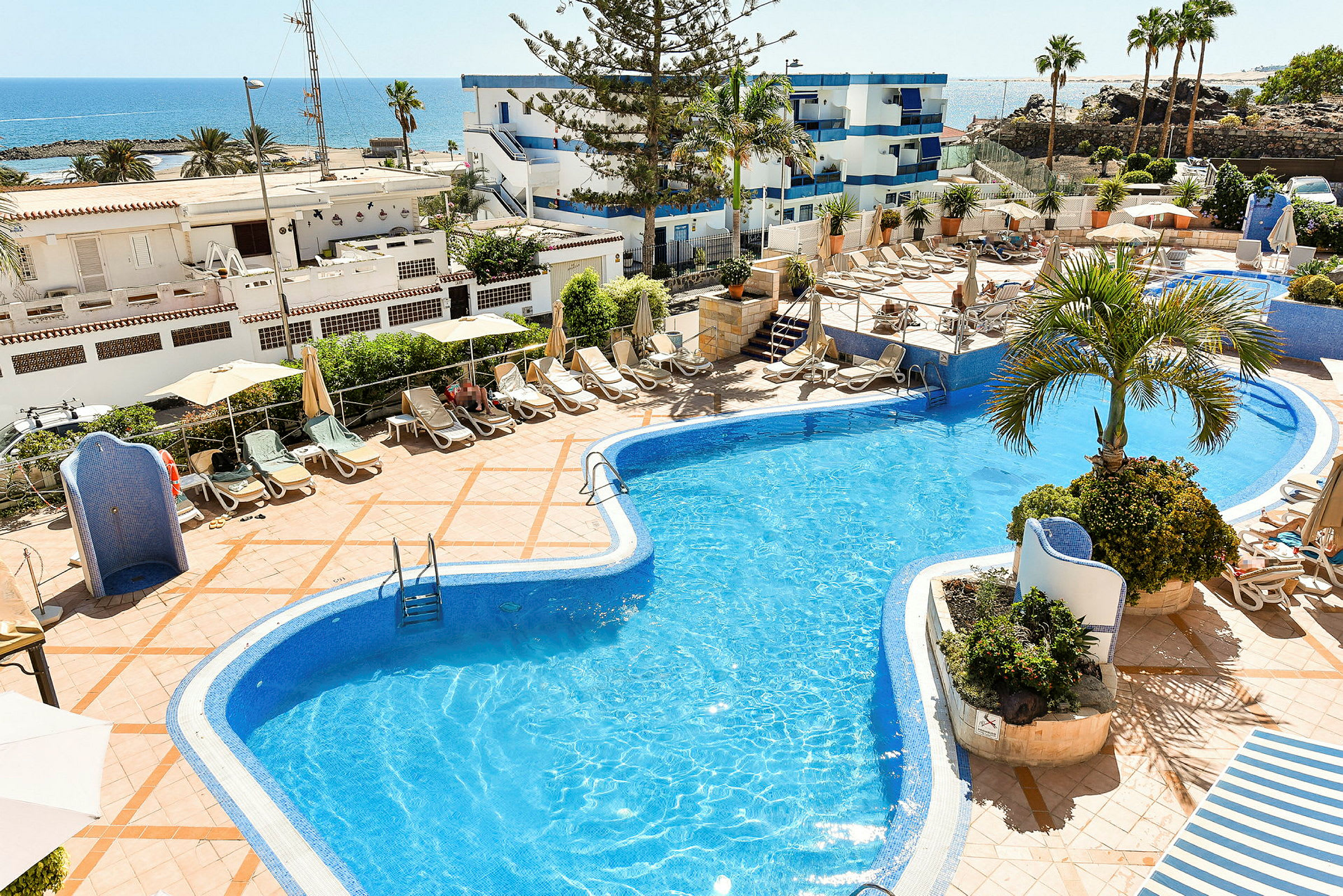 En pool med solstolar och parasoller på omgivande terrass. I bakgrunden syns havet och byggnader. (AI-genererad text)