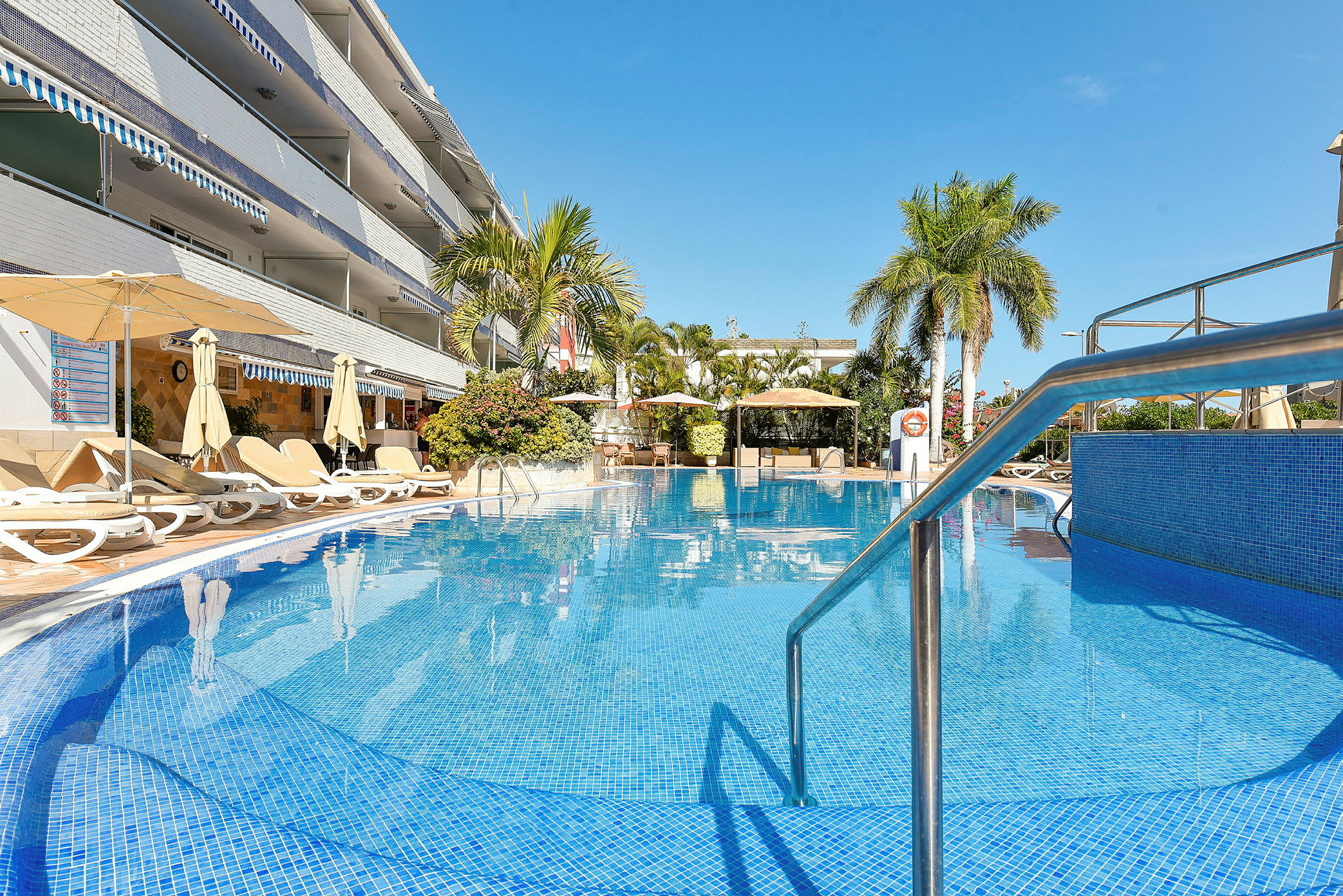 Utomhuspool vid hotell med solstolar och parasoller, omgiven av palmträd. (AI-genererad text)