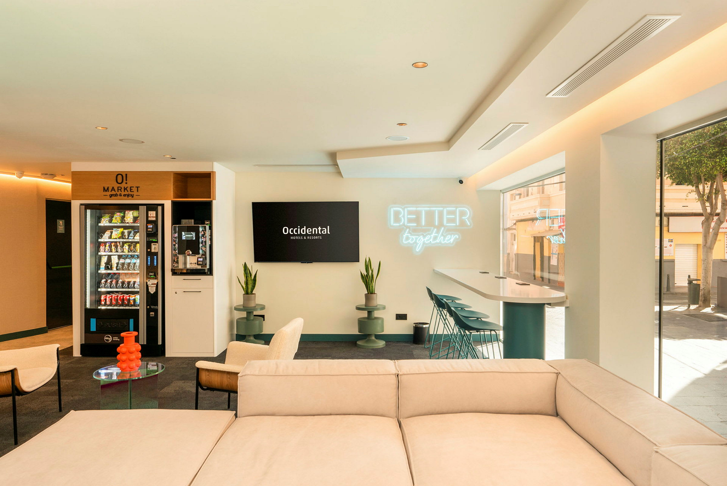 Ett modernt och ljust loungeområde med soffa, stolar, bord och barstolar vid ett högt bord. Väggen har en skylt med texten 'Better together' och en tv med logotypen för Occidental Hotels & Resorts. (AI-genererad text)