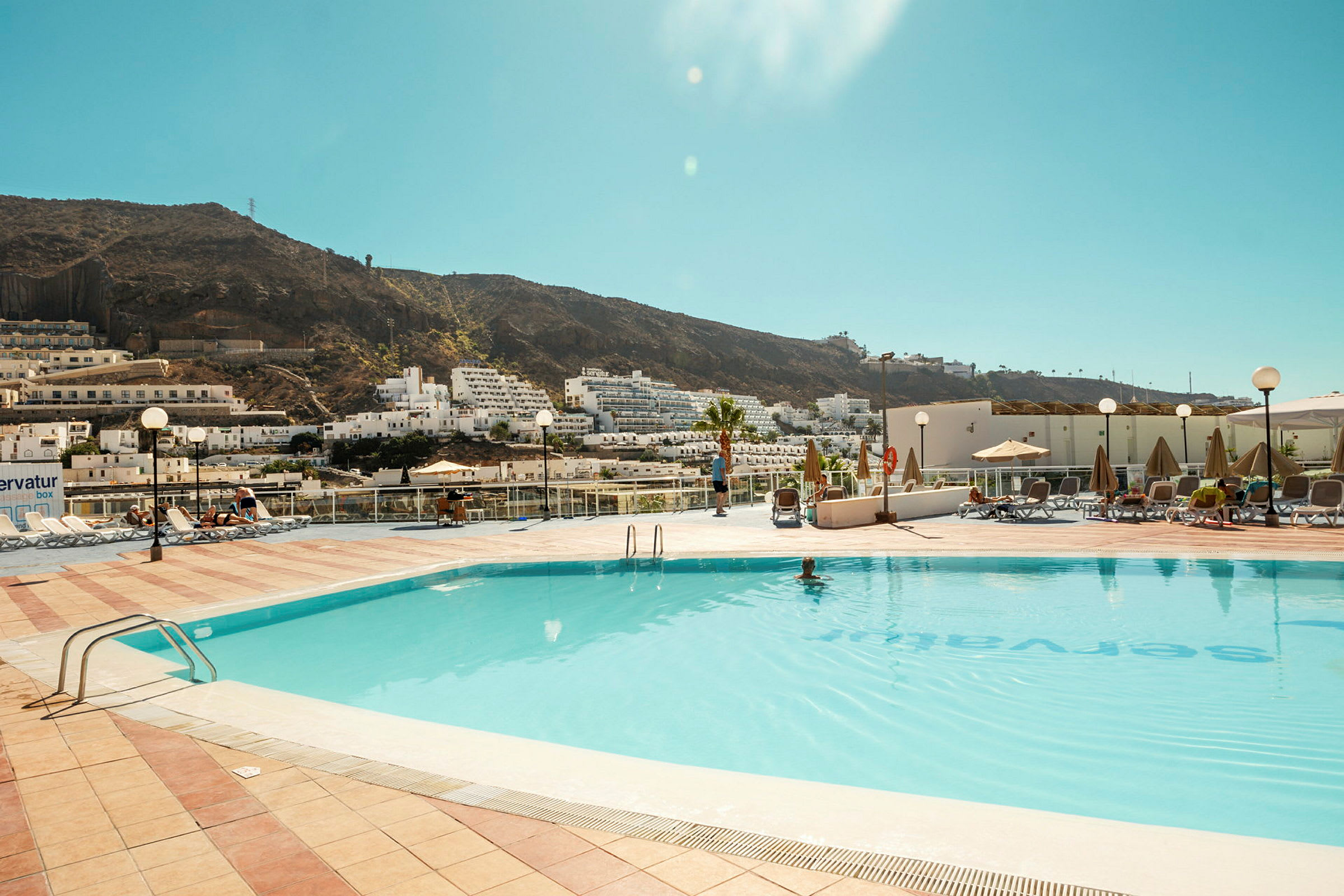 Poolområde med utsikt över en bergssluttning och vita byggnader. Solstolar och parasoller finns vid sidan. (AI-genererad text)